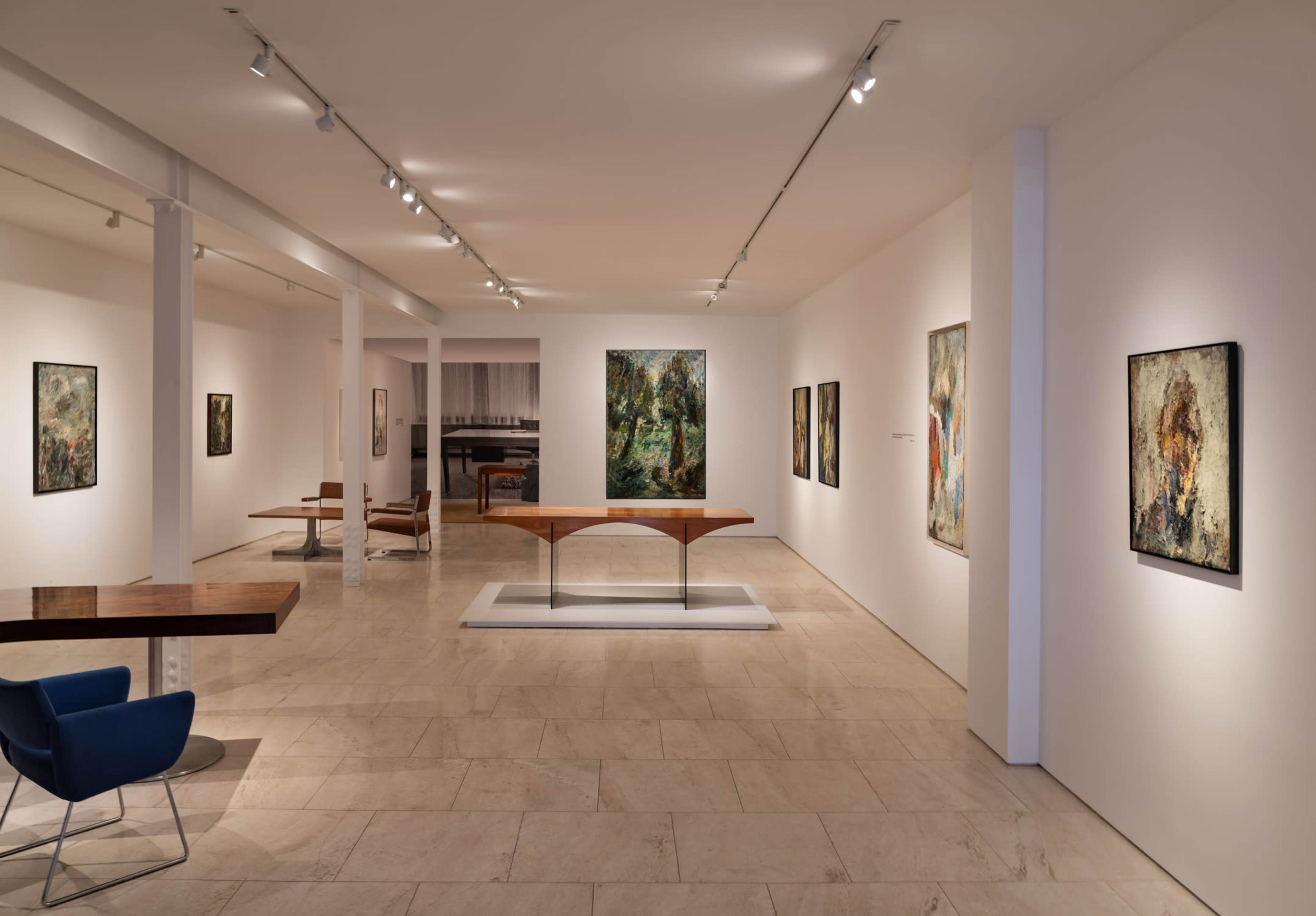 The image shows a modern art gallery featuring several framed paintings on white walls and a central wooden table with minimalist seating.
