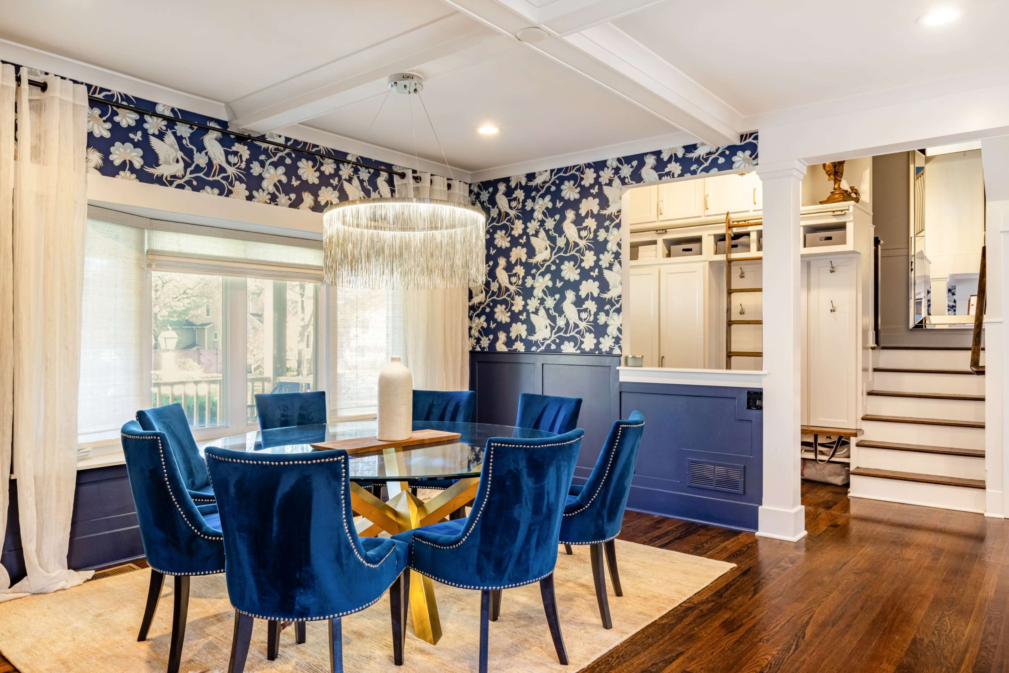 The image shows a dining room with a round glass table surrounded by six blue upholstered chairs, a large decorative chandelier, and floral wallpaper.