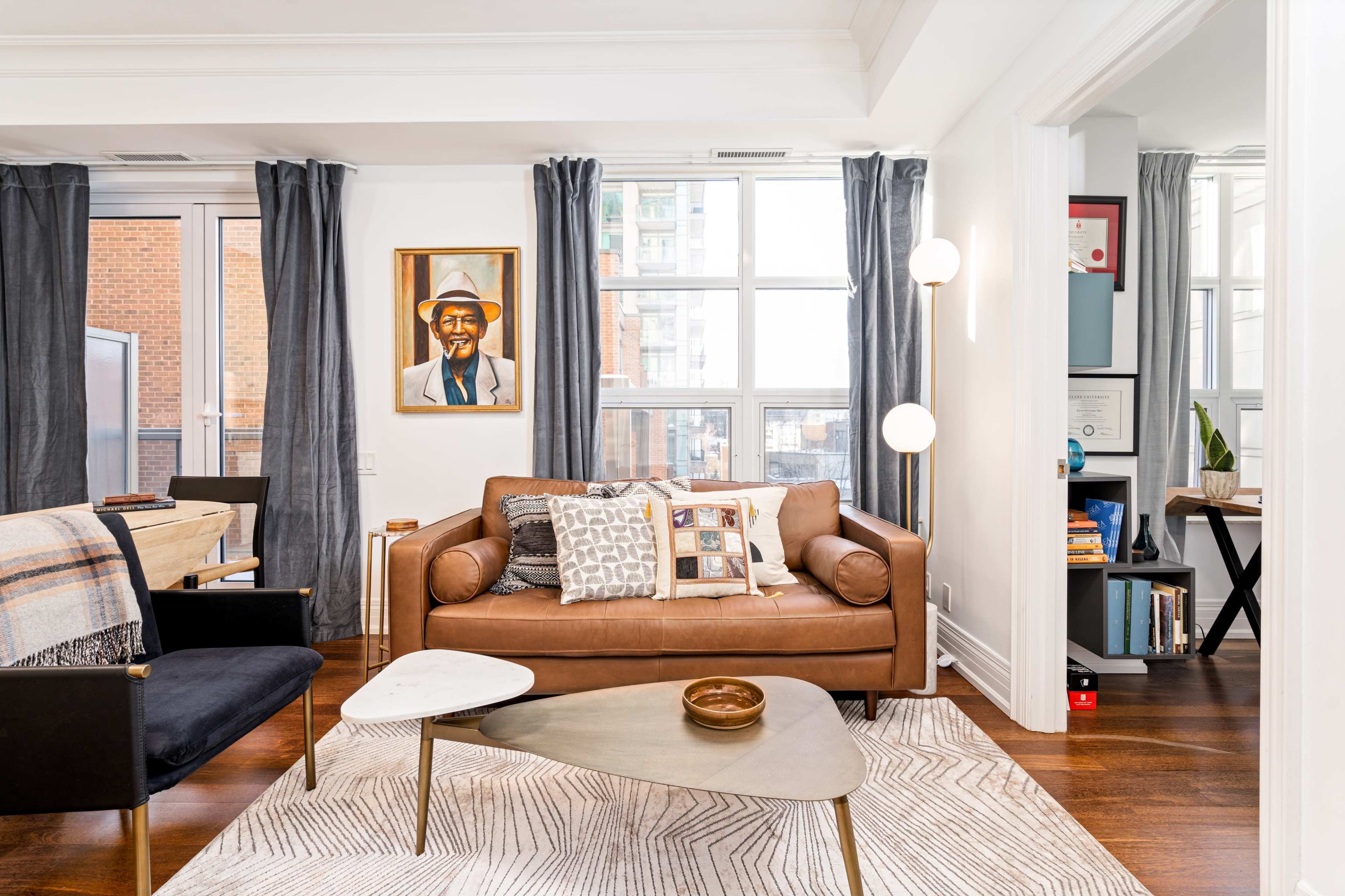 A modern living room with a brown leather sofa, a coffee table, and a decorative painting on the wall, illuminated by natural light from large windows.