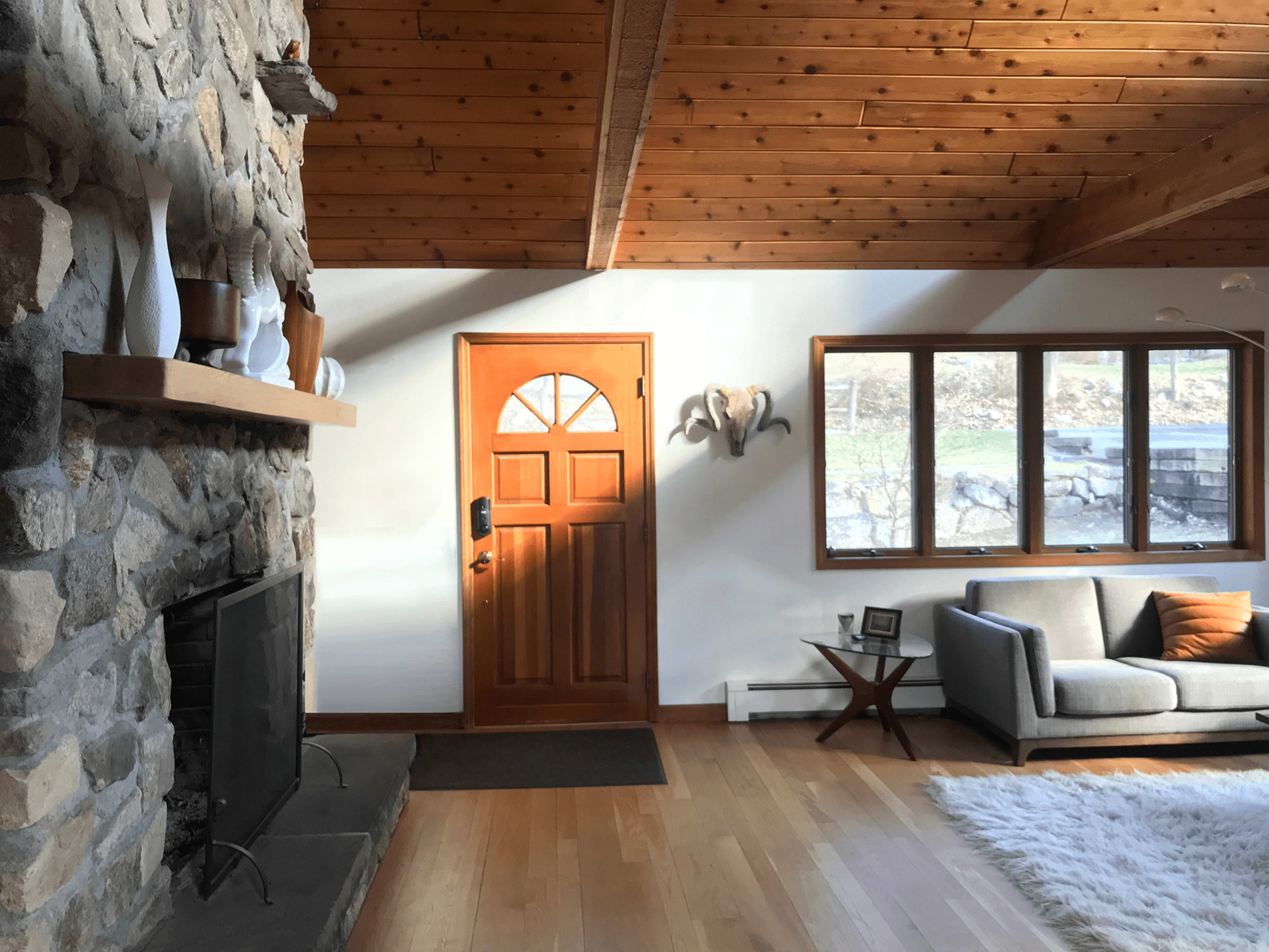 A stone fireplace is positioned next to a wooden door, with a large window and a gray sofa in a sunlit living room.