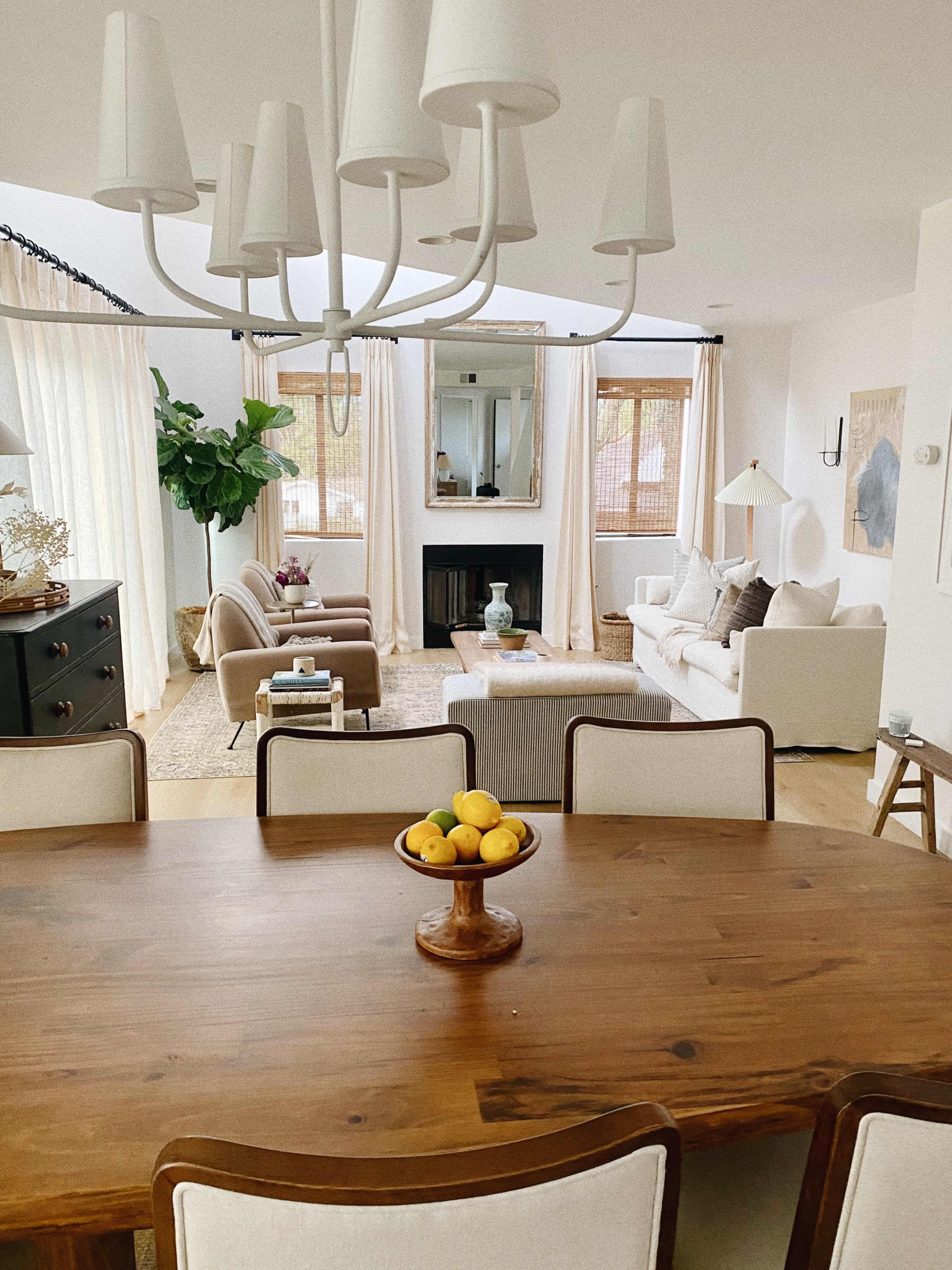 A dining table with a bowl of lemons is set in the foreground, facing a bright living room with two armchairs, a sofa, and large windows.