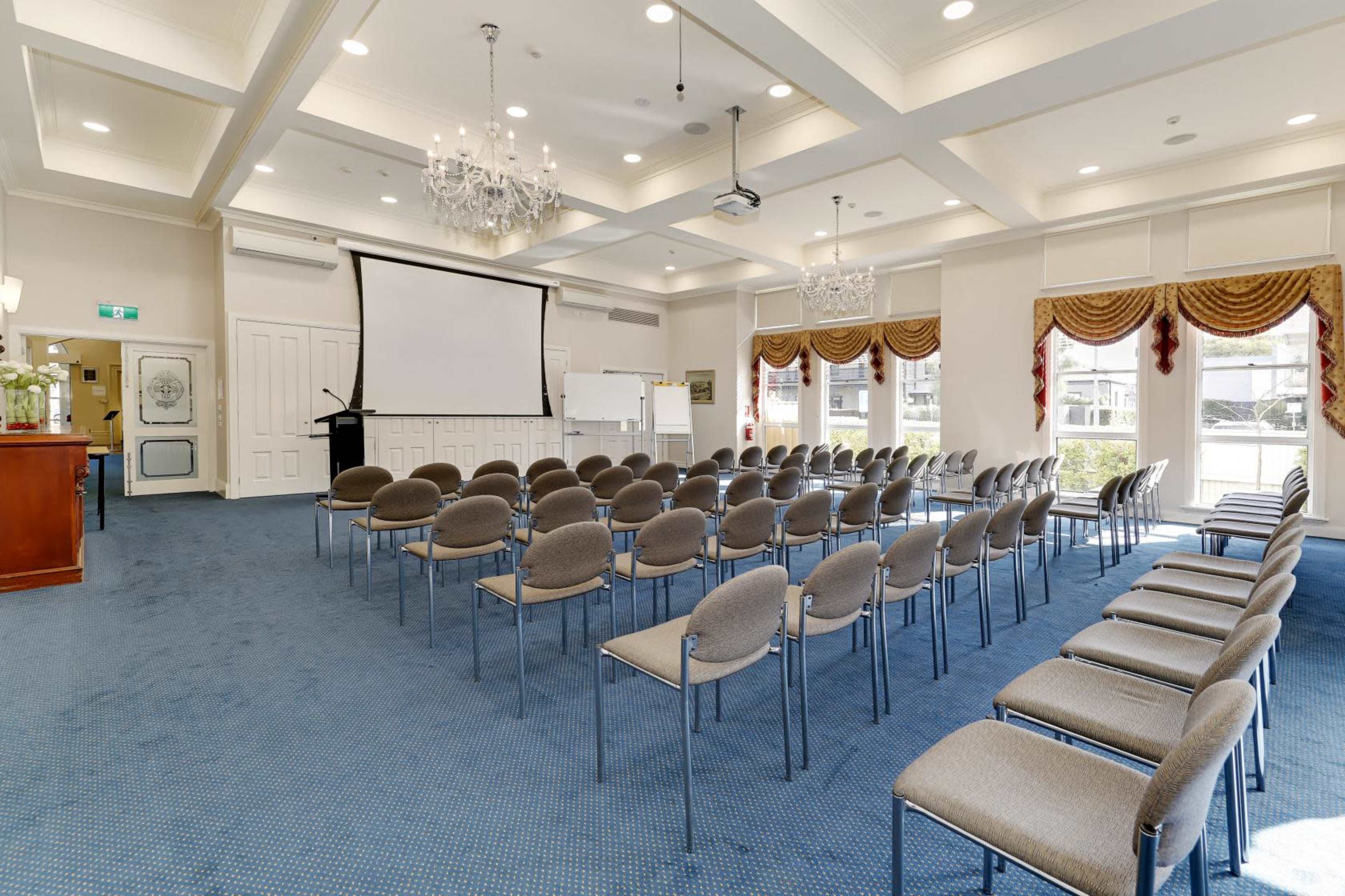 A conference room with rows of chairs facing a projection screen and a podium, featuring a carpeted floor and decorative chandeliers.