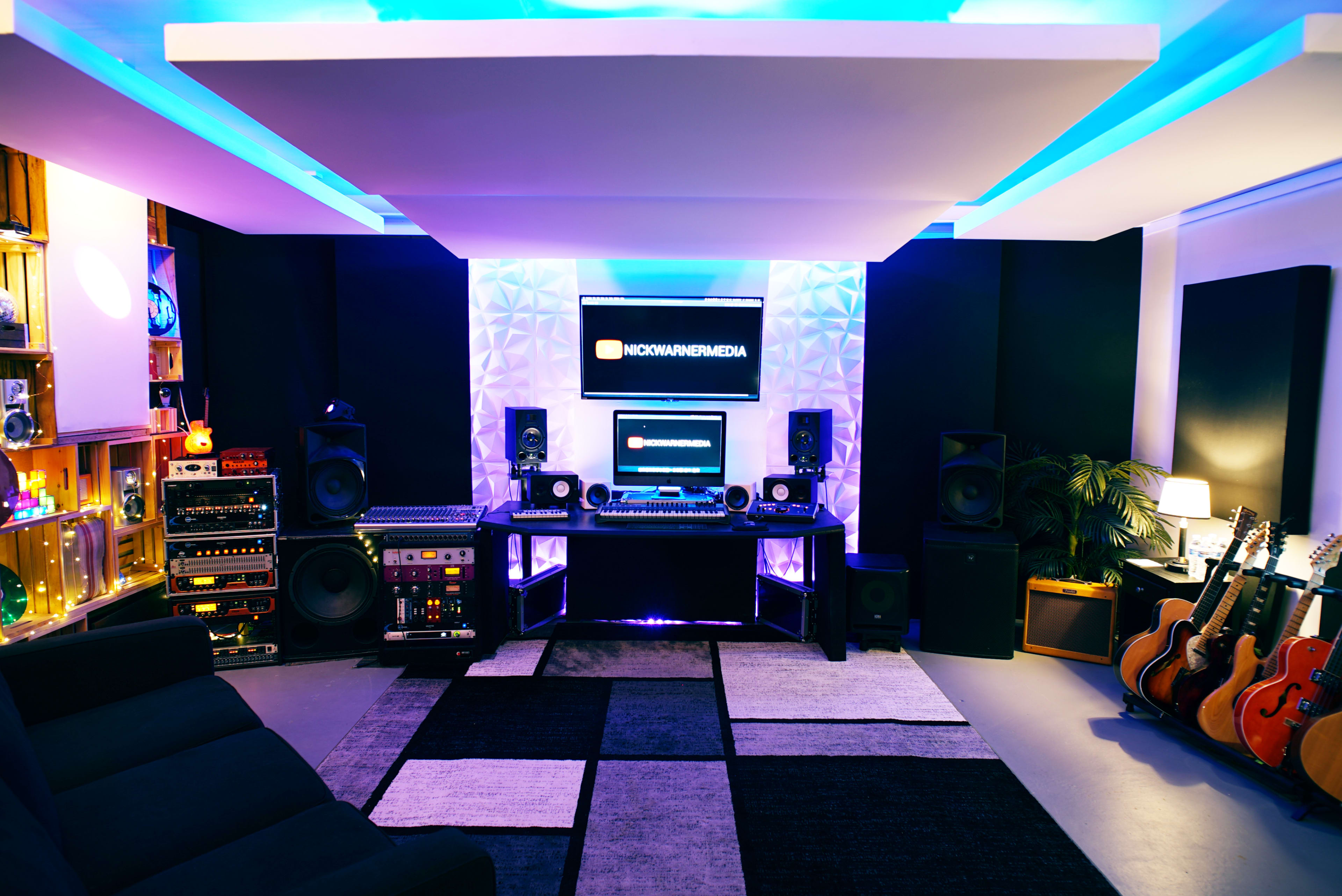 A modern recording studio features a central desk with audio equipment, surrounded by speakers and guitars, bathed in blue and purple lighting.