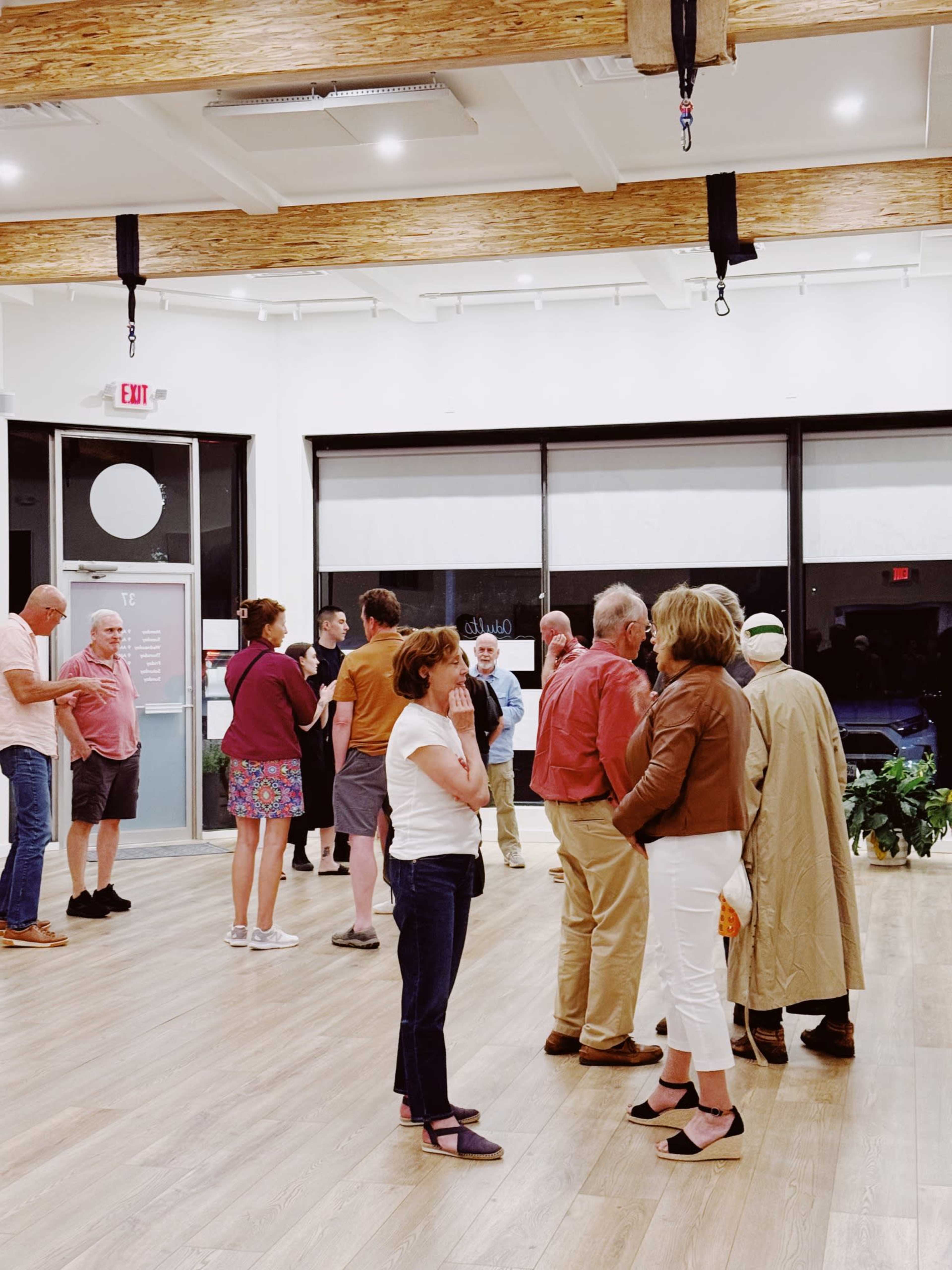 A group of people chat and mingle in a spacious interior with wooden beams and large windows.