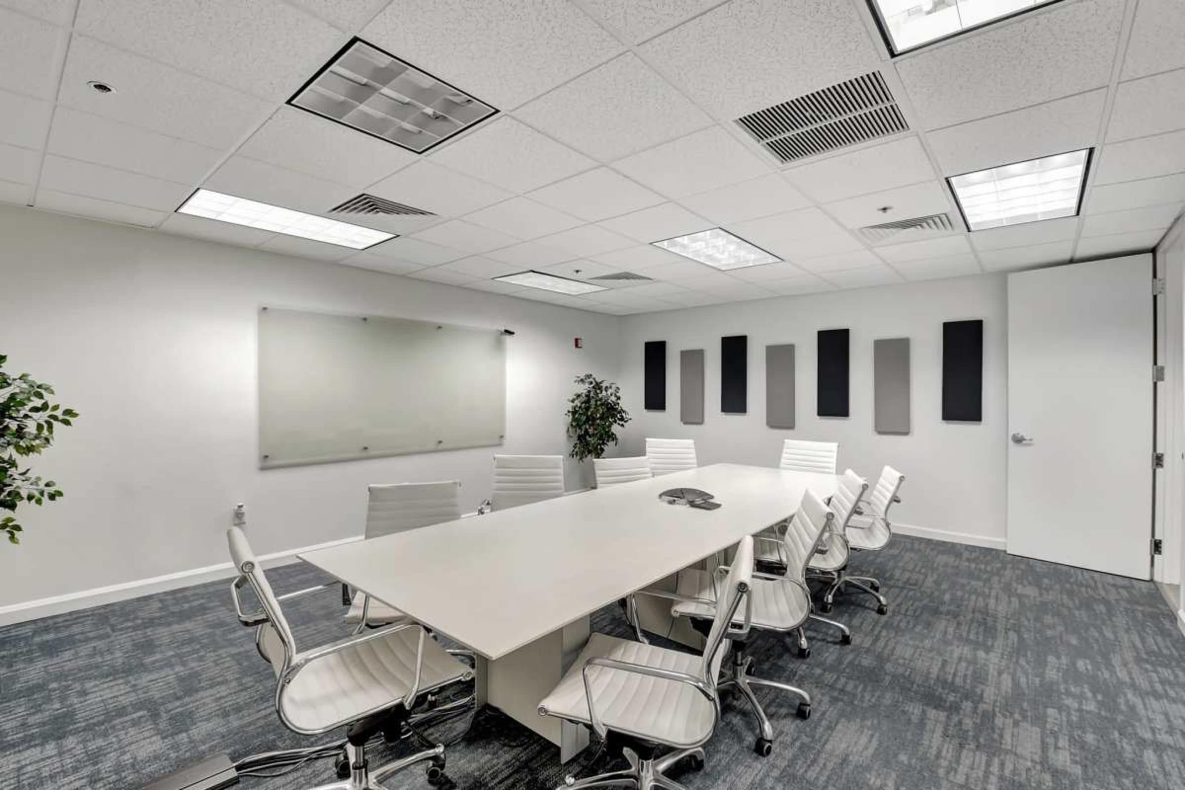 A modern conference room features a long table surrounded by white chairs, with a large whiteboard and abstract wall panels.