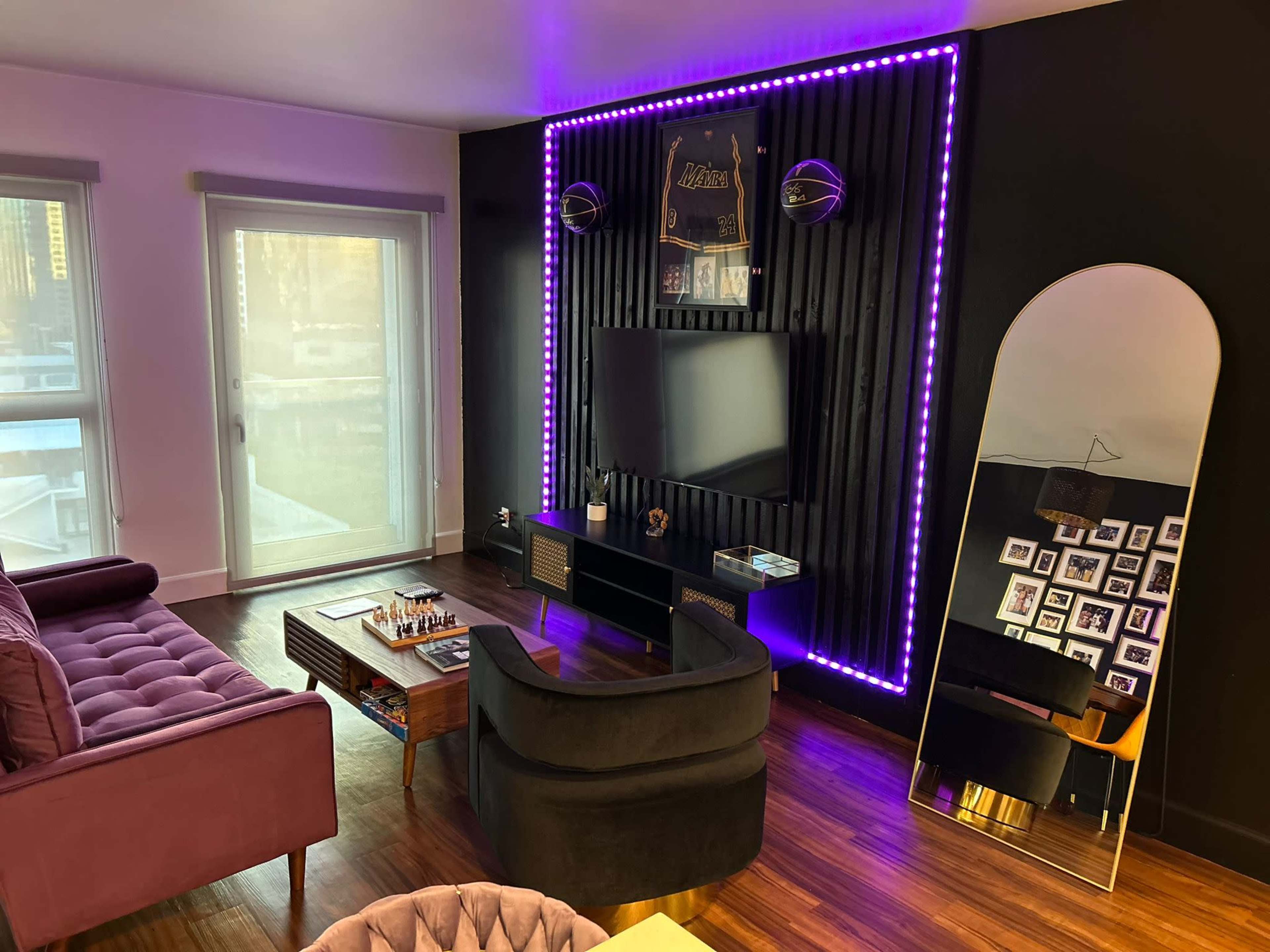 The image features a modern living room with a purple couch, a black TV stand, an illuminated wall, and a large mirror beside a collection of framed photos.