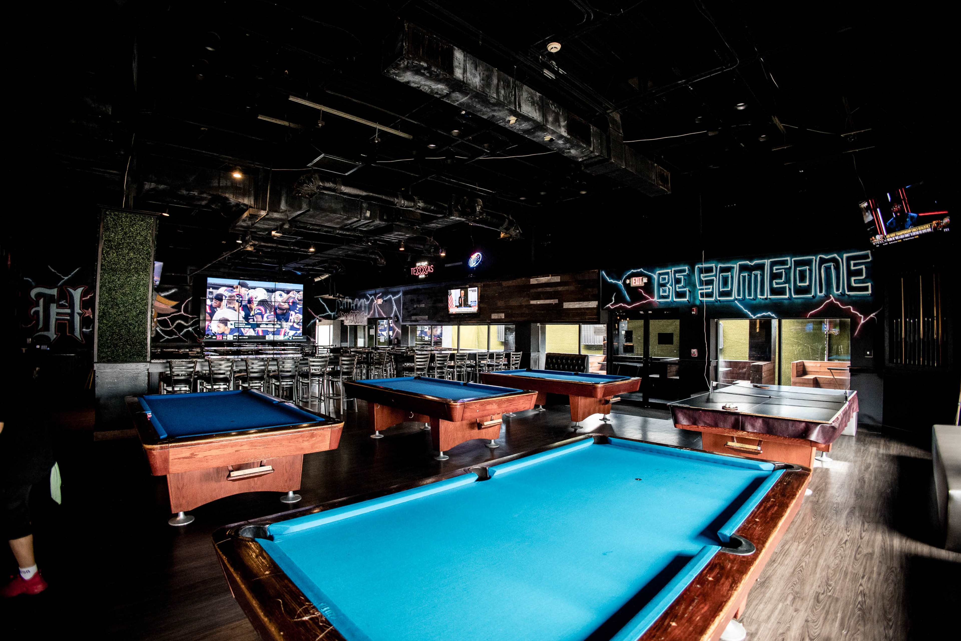 The image shows a spacious pool hall with several blue billiard tables and a large screen displaying a game, alongside a bar area in the background.