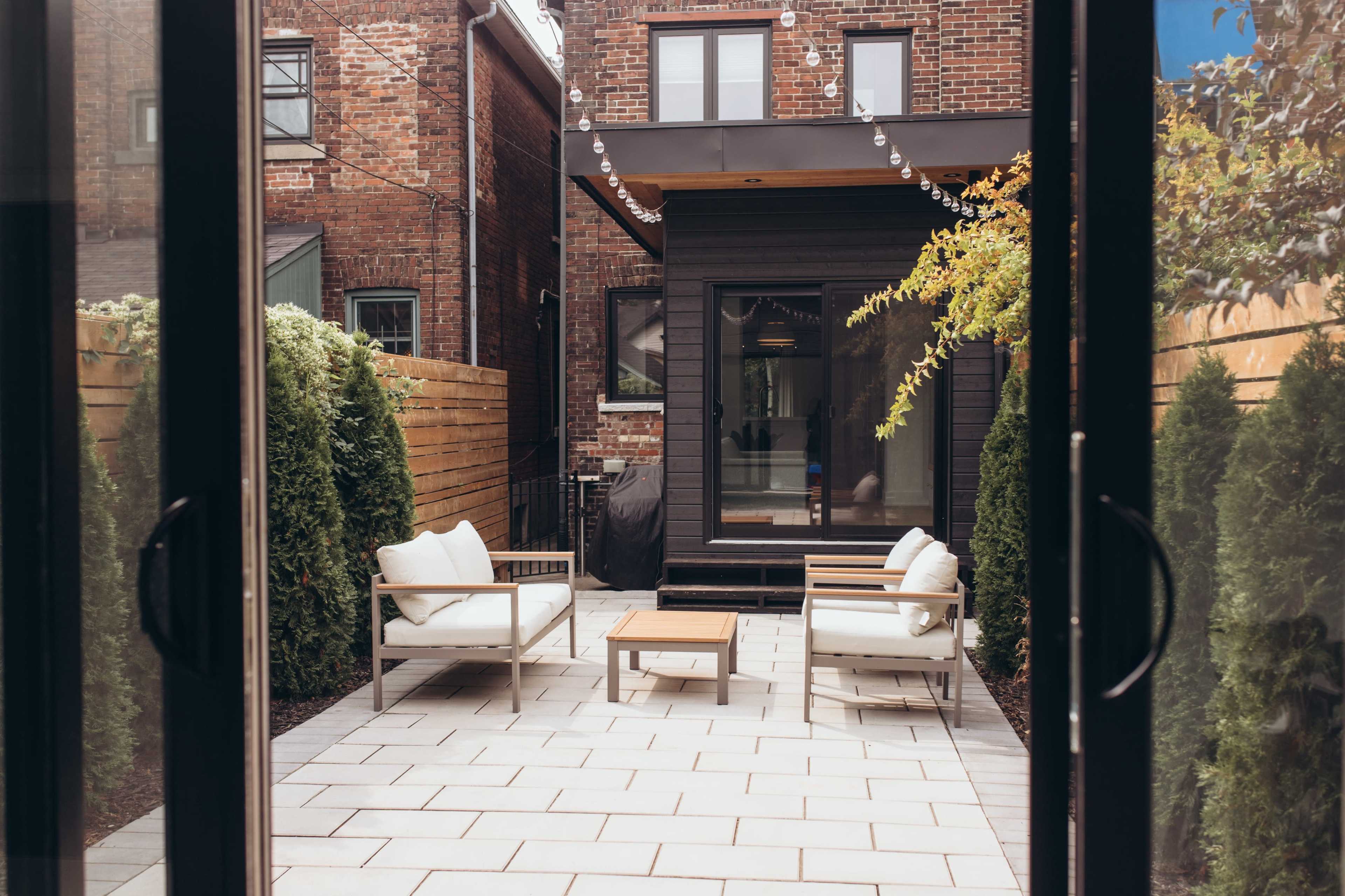A small outdoor patio features two chairs and a table, surrounded by greenery and brick buildings.