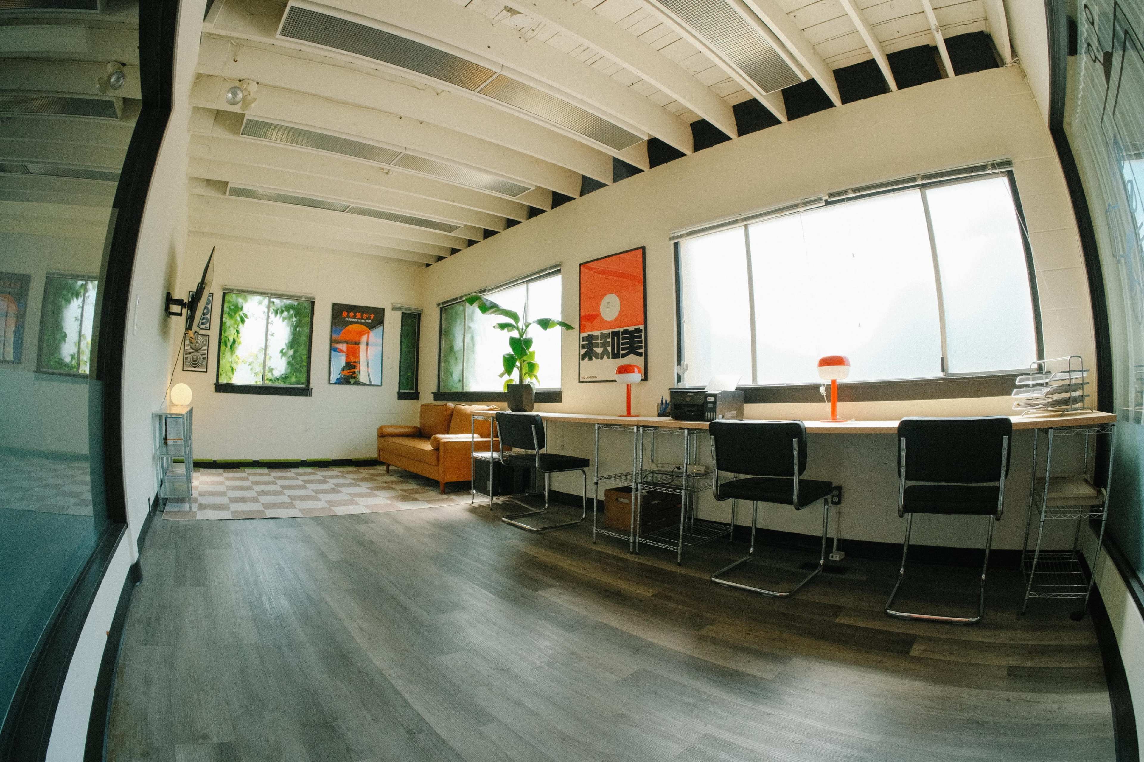 The image shows a modern office space with a sofa, a desk with chairs, and large windows letting in natural light.