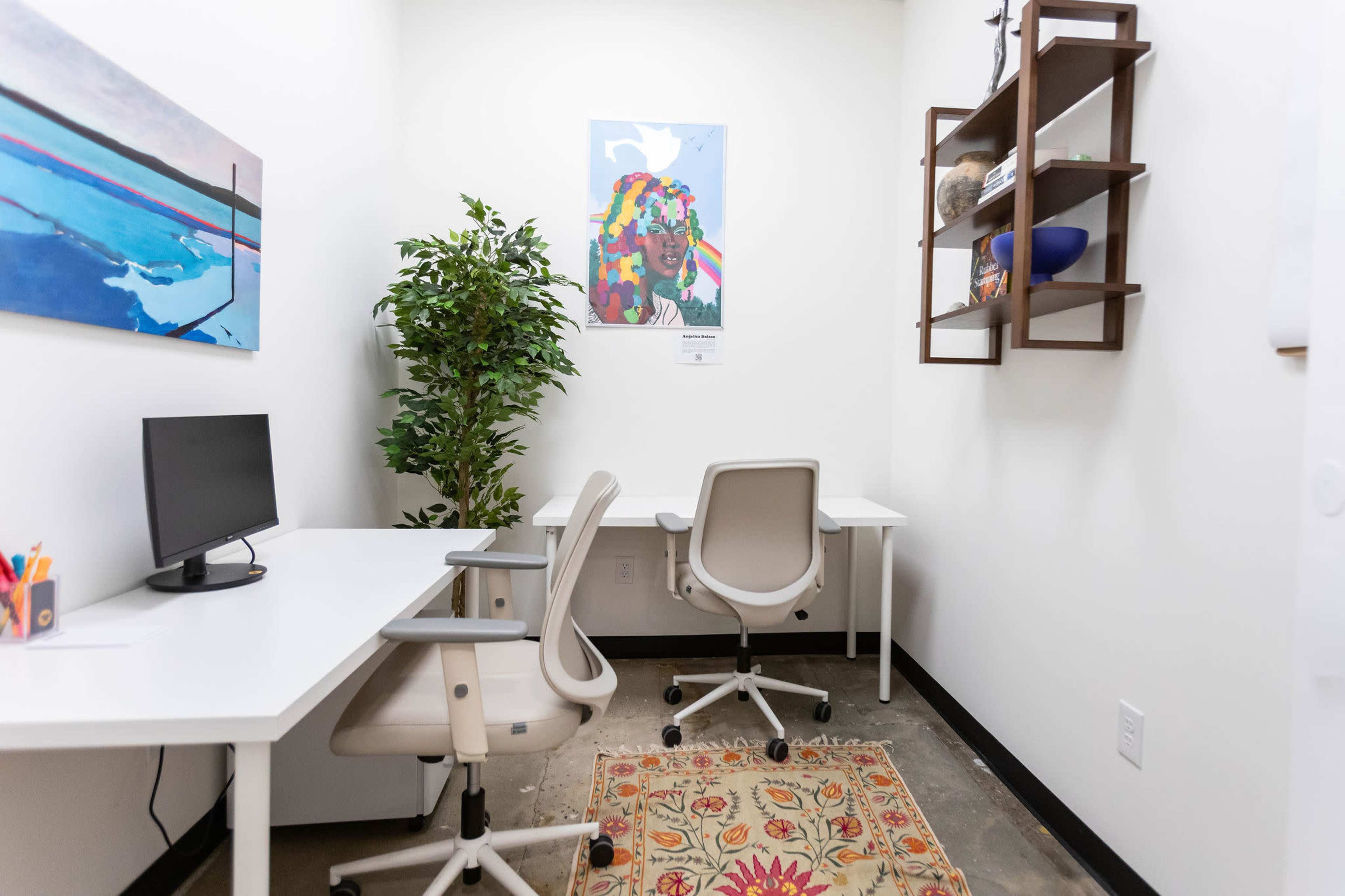 The image shows a small office space with two desks, each equipped with a chair, a potted plant, and a colorful artwork hanging on the wall.