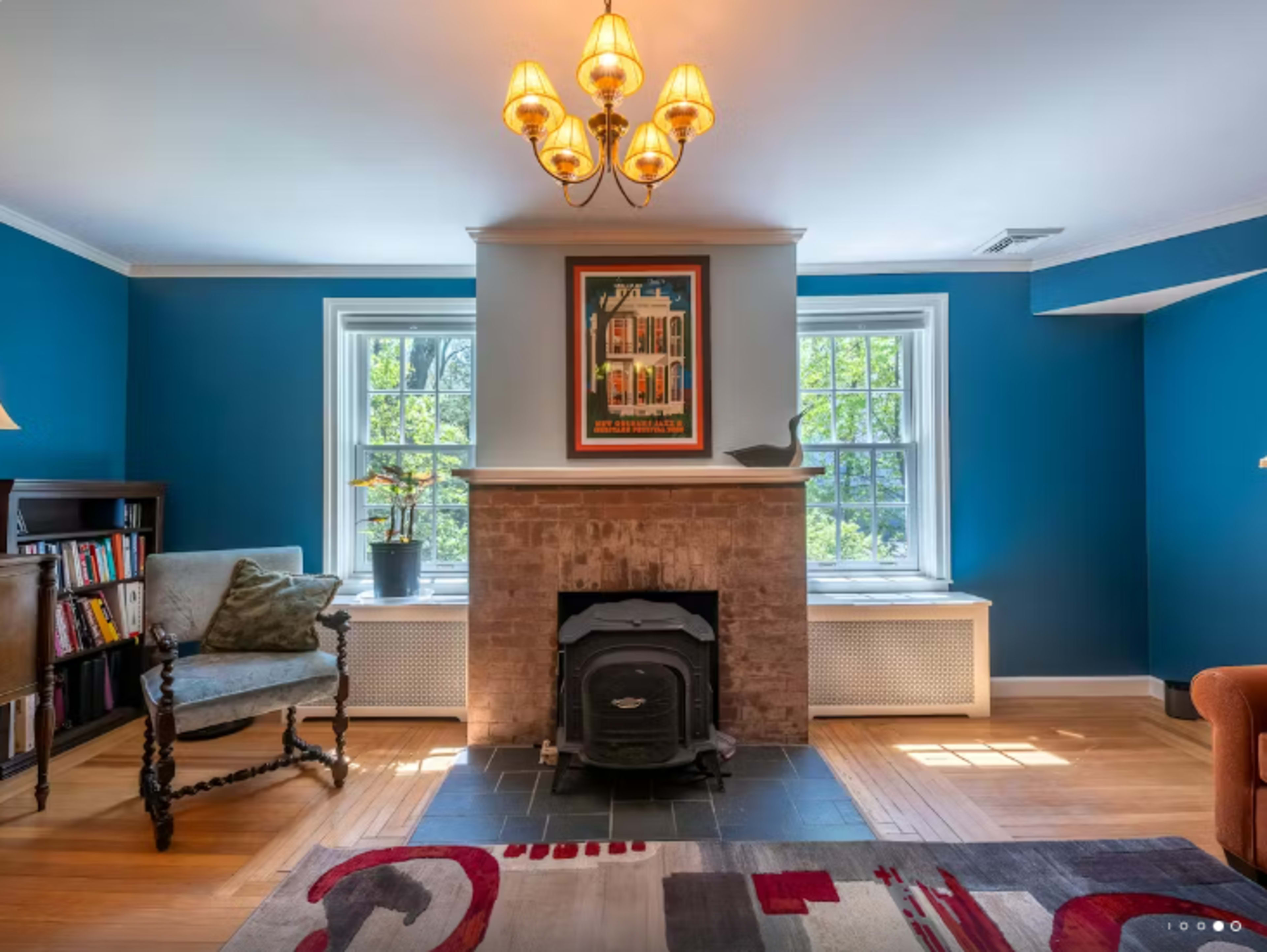 The image shows a cozy living room featuring a brick fireplace, blue walls, a wooden chair, and a colorful rug.