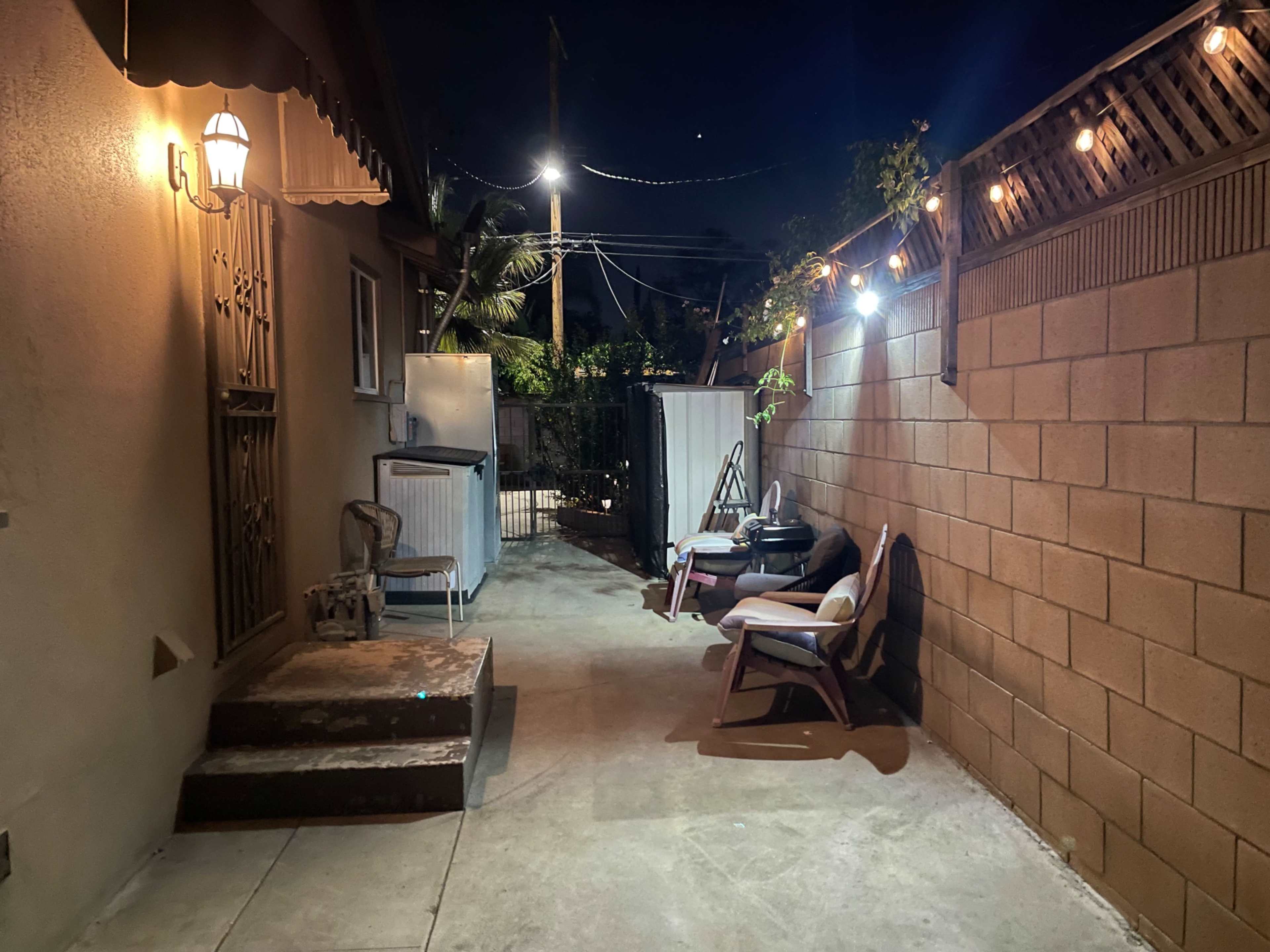 The scene depicts a narrow, well-lit alleyway beside a building, featuring a few chairs, a set of steps, and decorative string lights overhead.