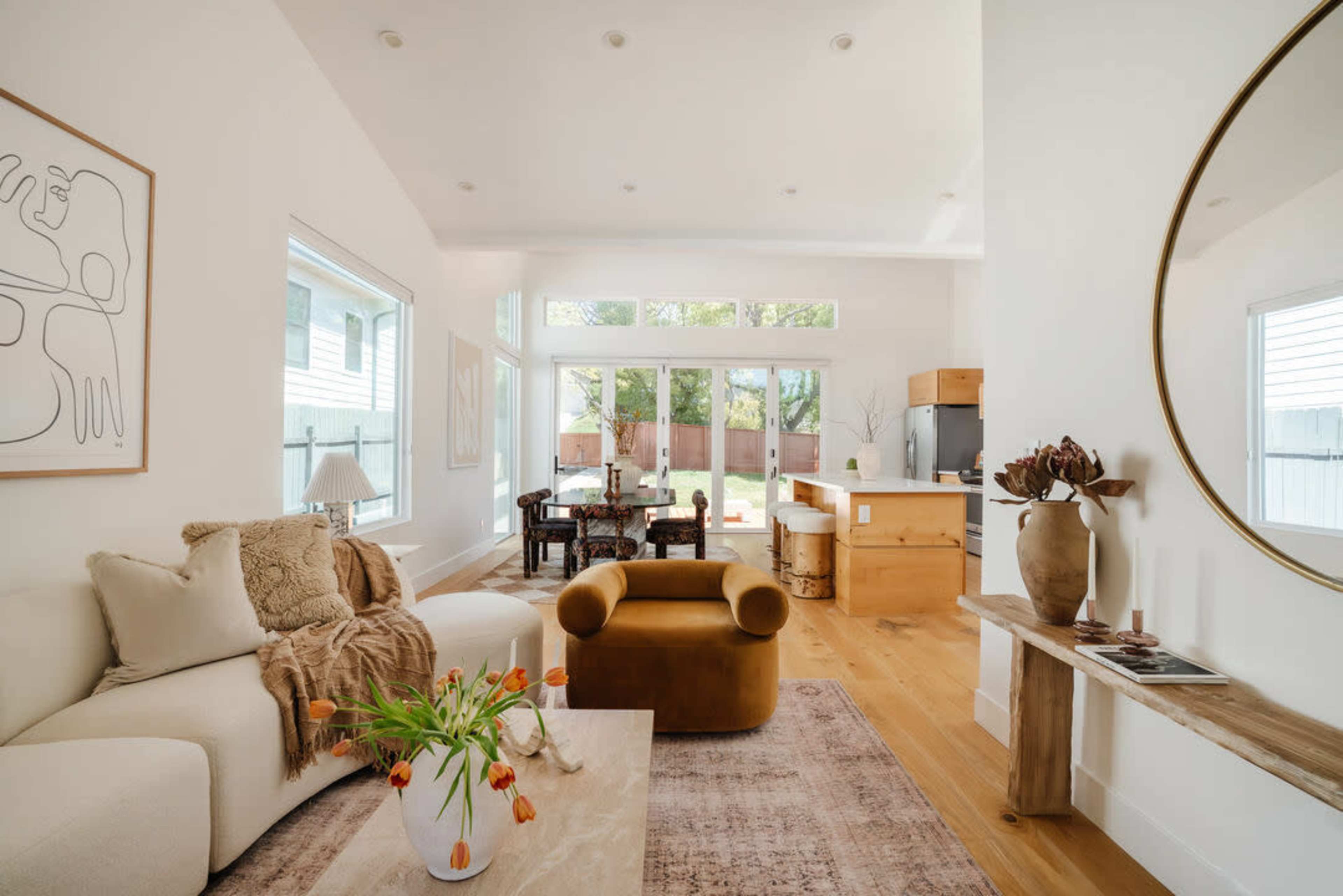 The image shows a modern living room with a light color palette, featuring a curved sofa, a coffee table with flowers, and a view of an outdoor area through large windows.