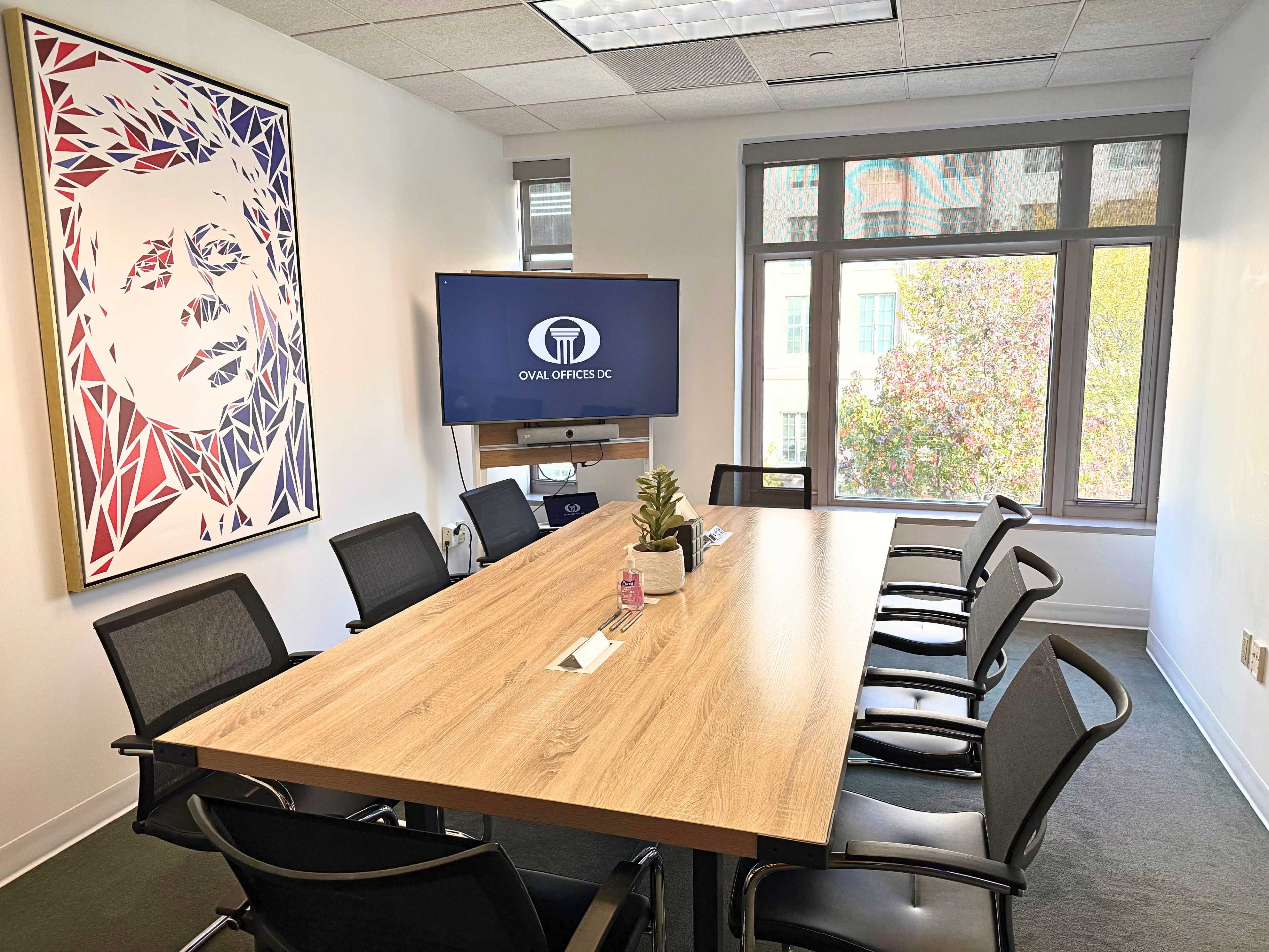 The image shows a modern conference room with a large wooden table, several black chairs, a wall-mounted TV displaying a logo, and a large artwork of a person on the wall.