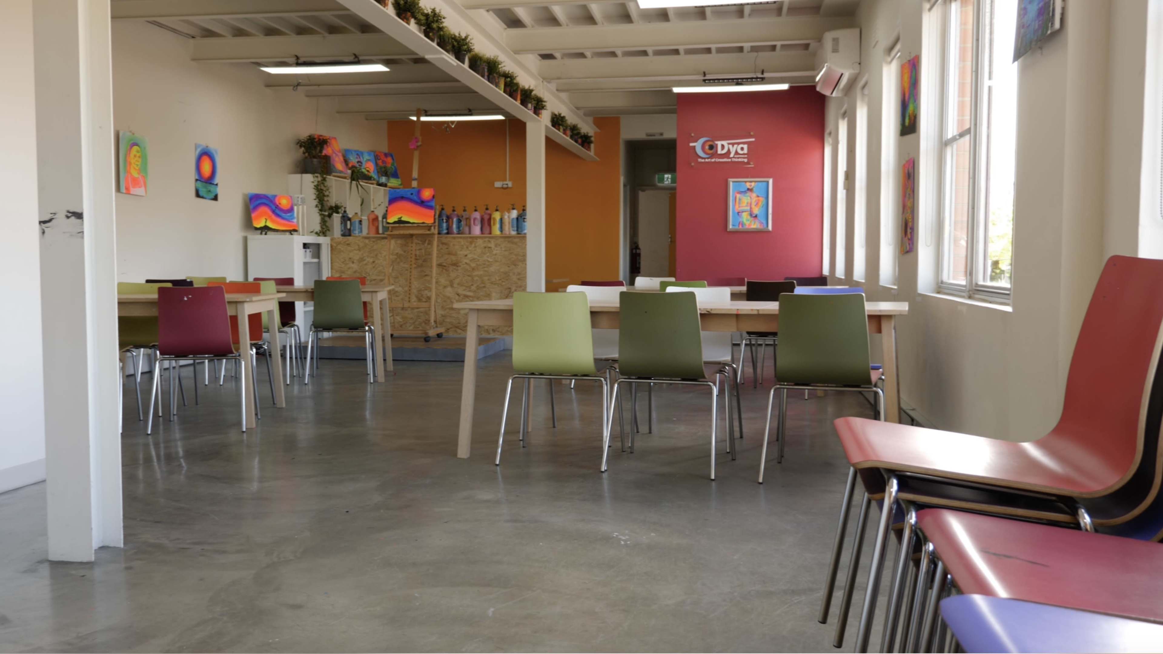 A colorful art studio features tables and chairs arranged around a spacious room with artwork displayed on the walls.