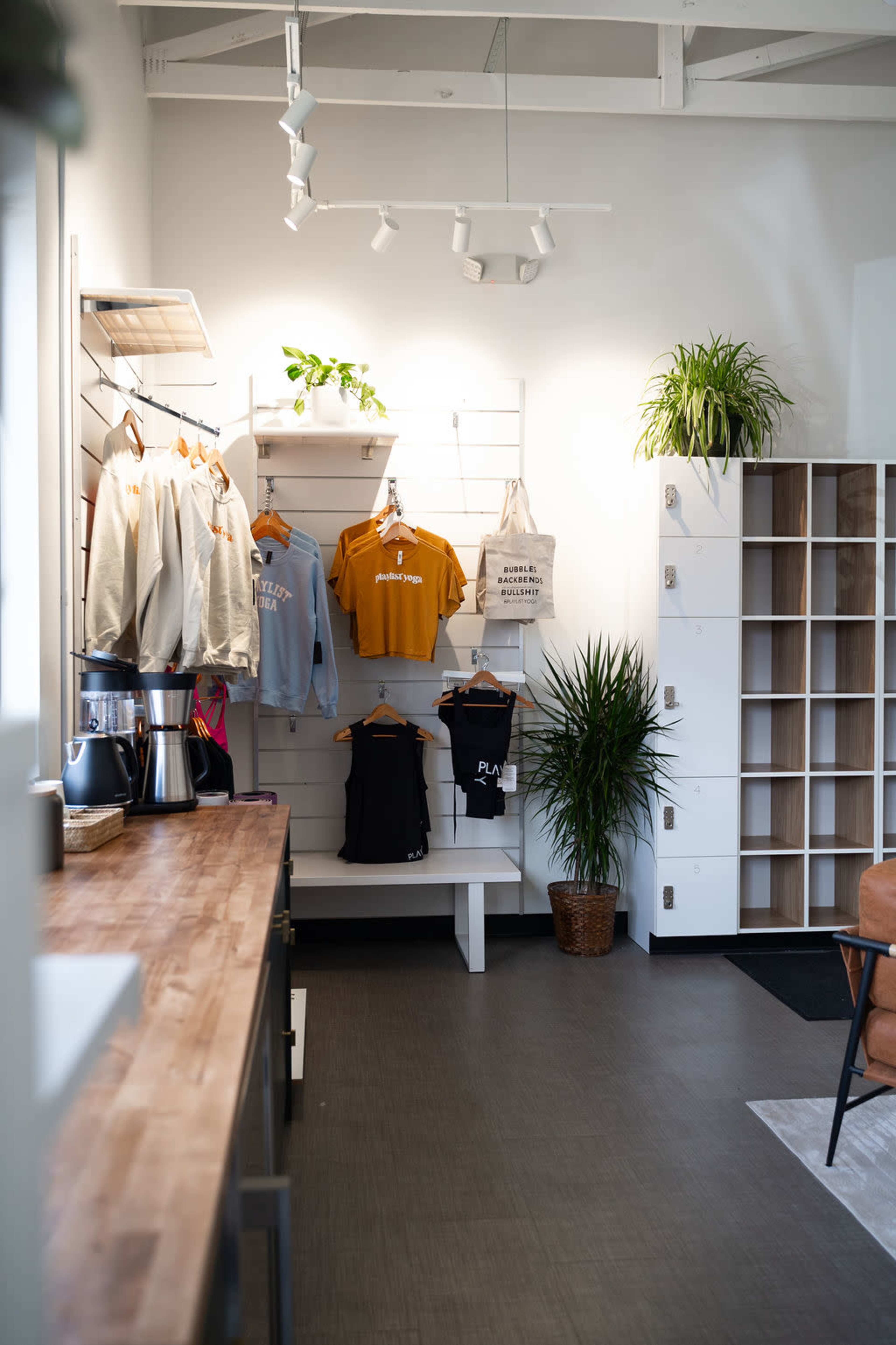 The image shows an interior of a retail space featuring clothing displays, a wooden counter, plants, and storage units.
