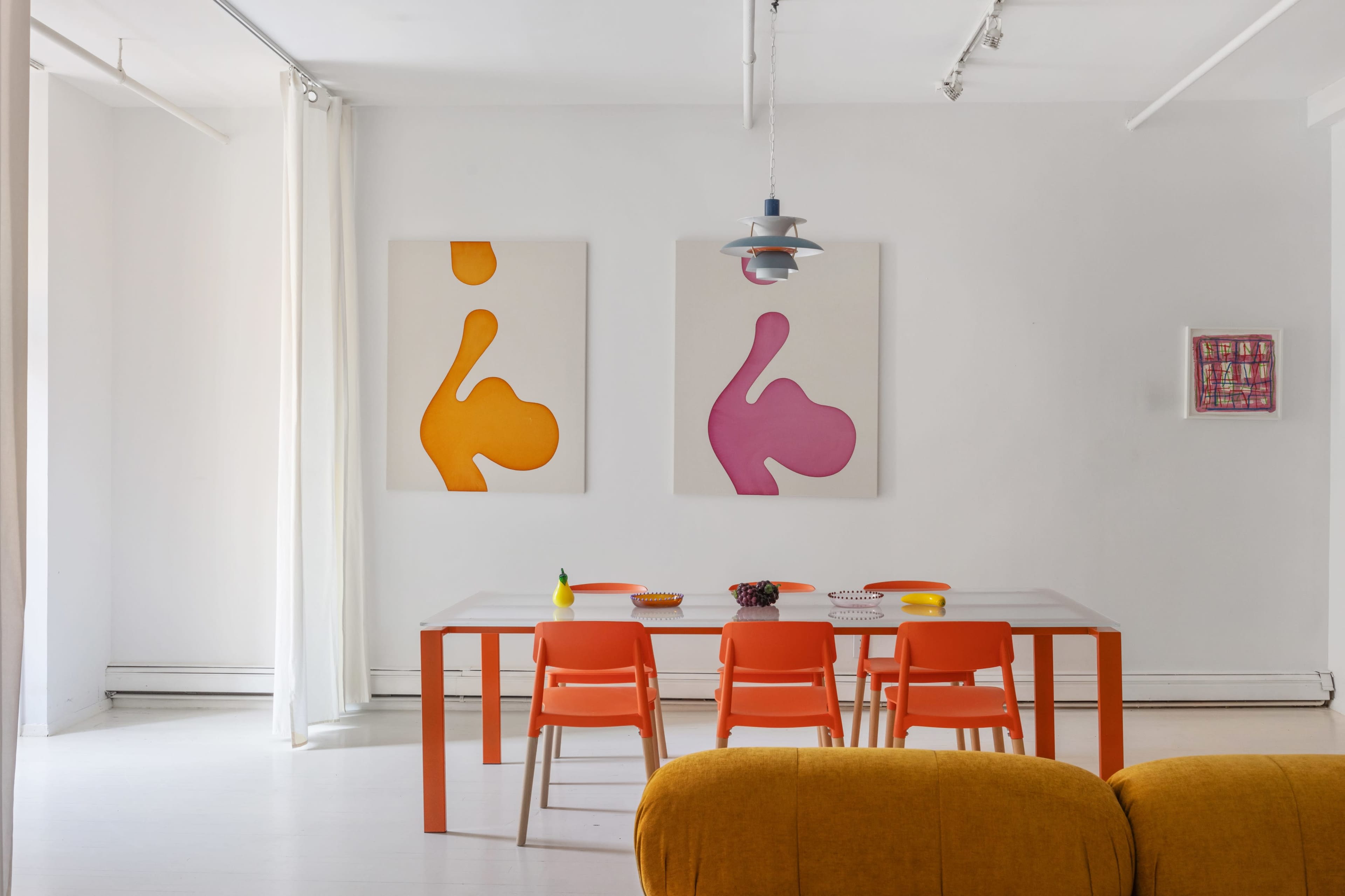 The image shows a bright dining area with a white table surrounded by orange chairs, displaying colorful plates, and featuring abstract art on the walls.