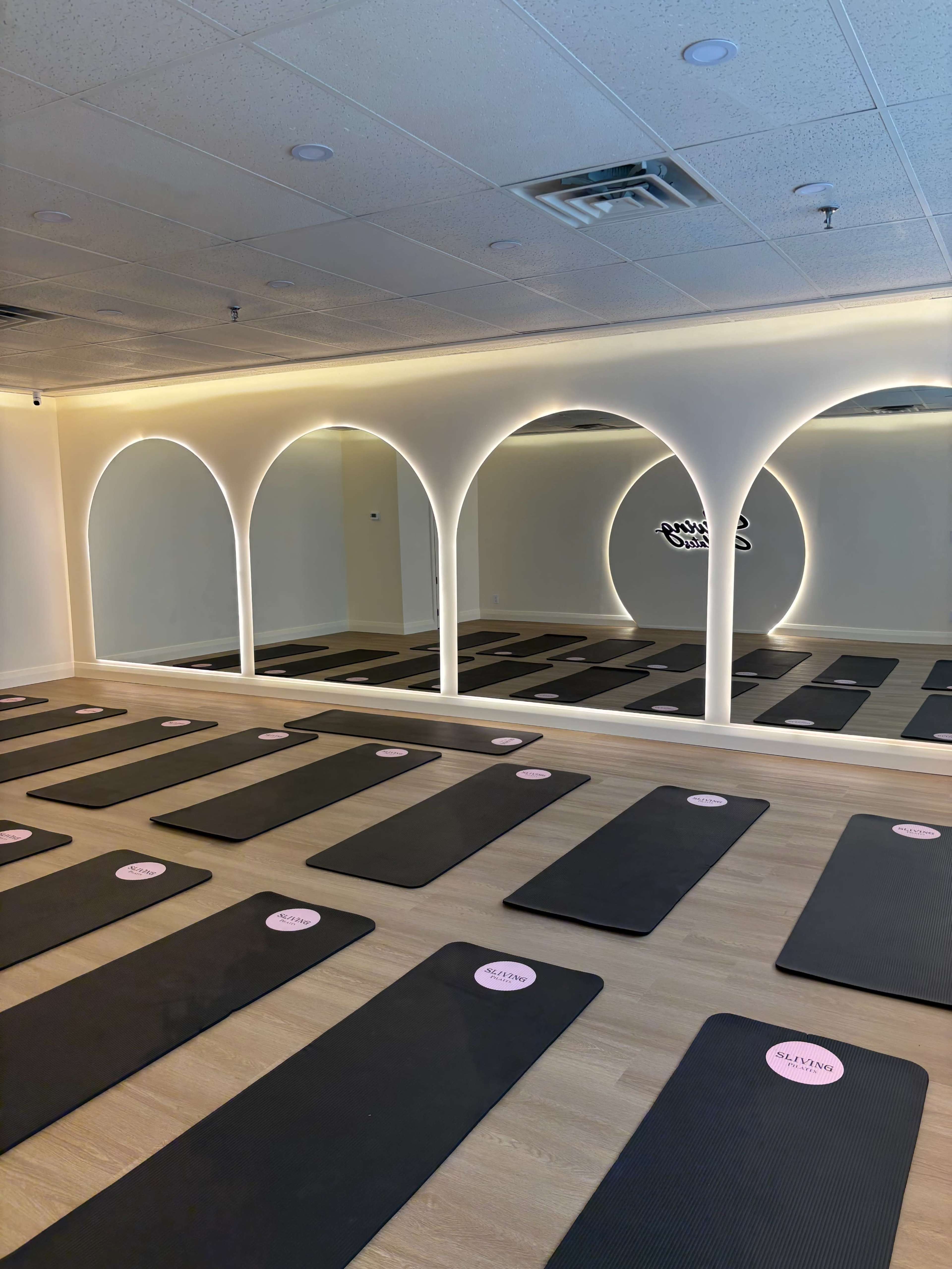 The image shows a spacious yoga studio with neatly arranged black mats on a wooden floor, illuminated by soft lighting and featuring a mirrored wall with arched frames.