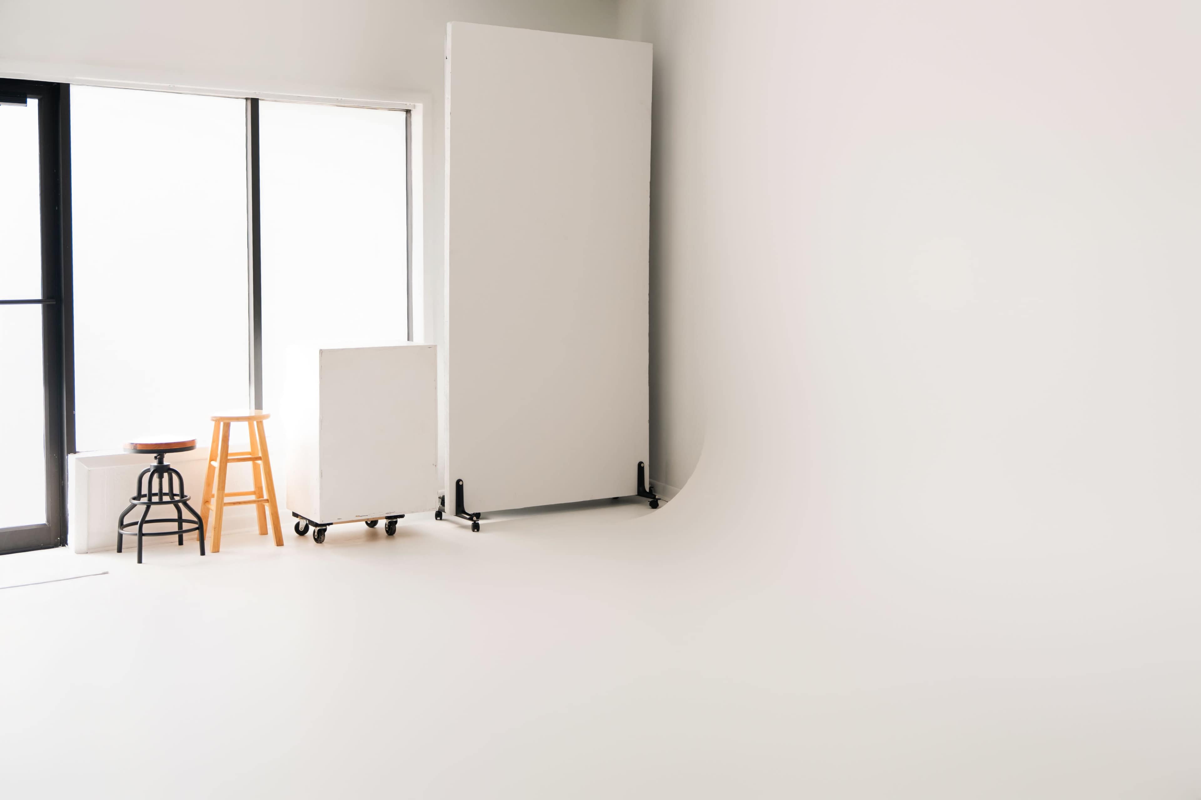 The image shows a minimalist studio space featuring a large white backdrop, a stool, and a small cart against a large window.