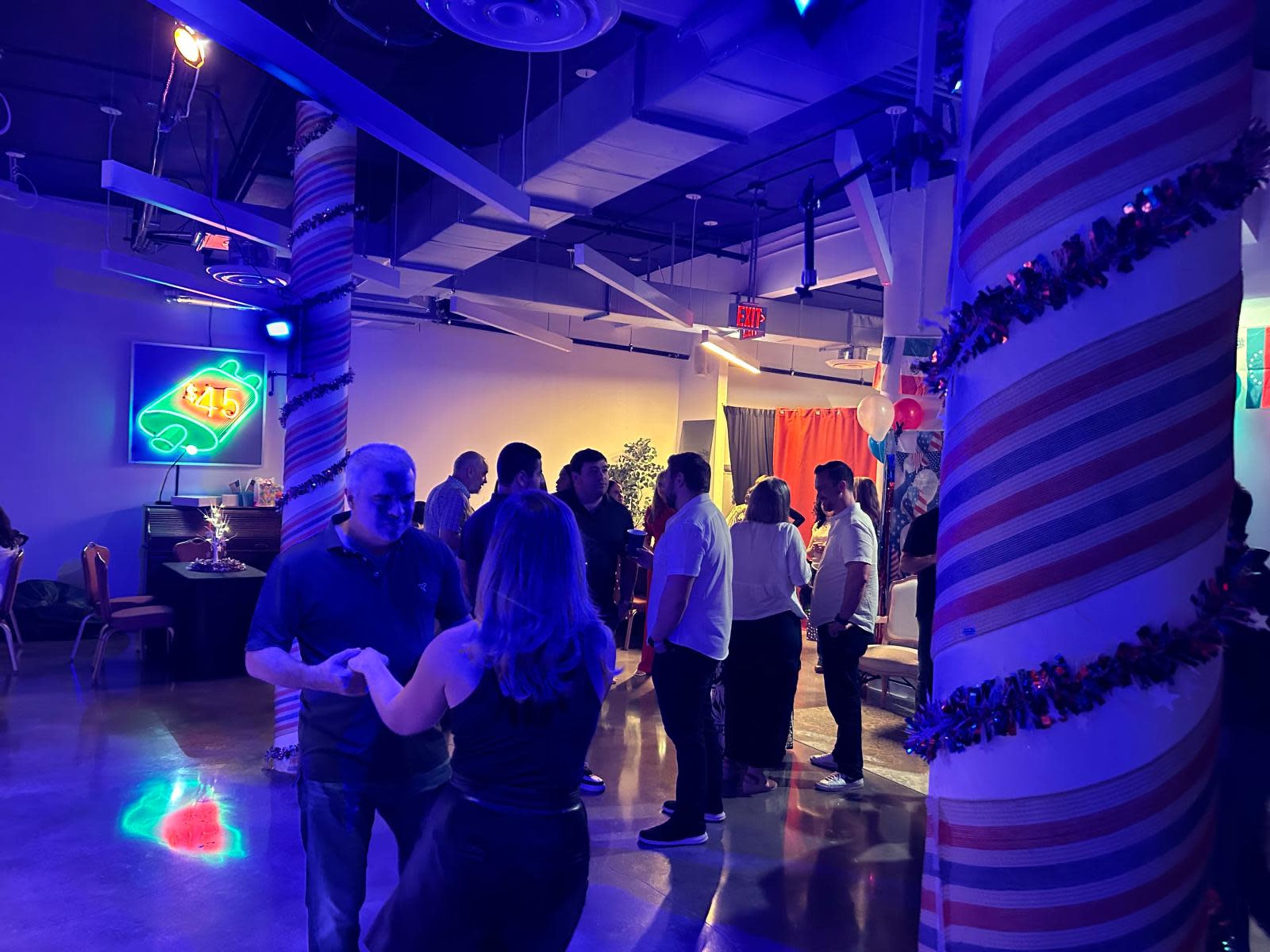 The image shows a lively indoor event with people socializing, dancing, and a colorful decor featuring illuminated signs and festive decorations.