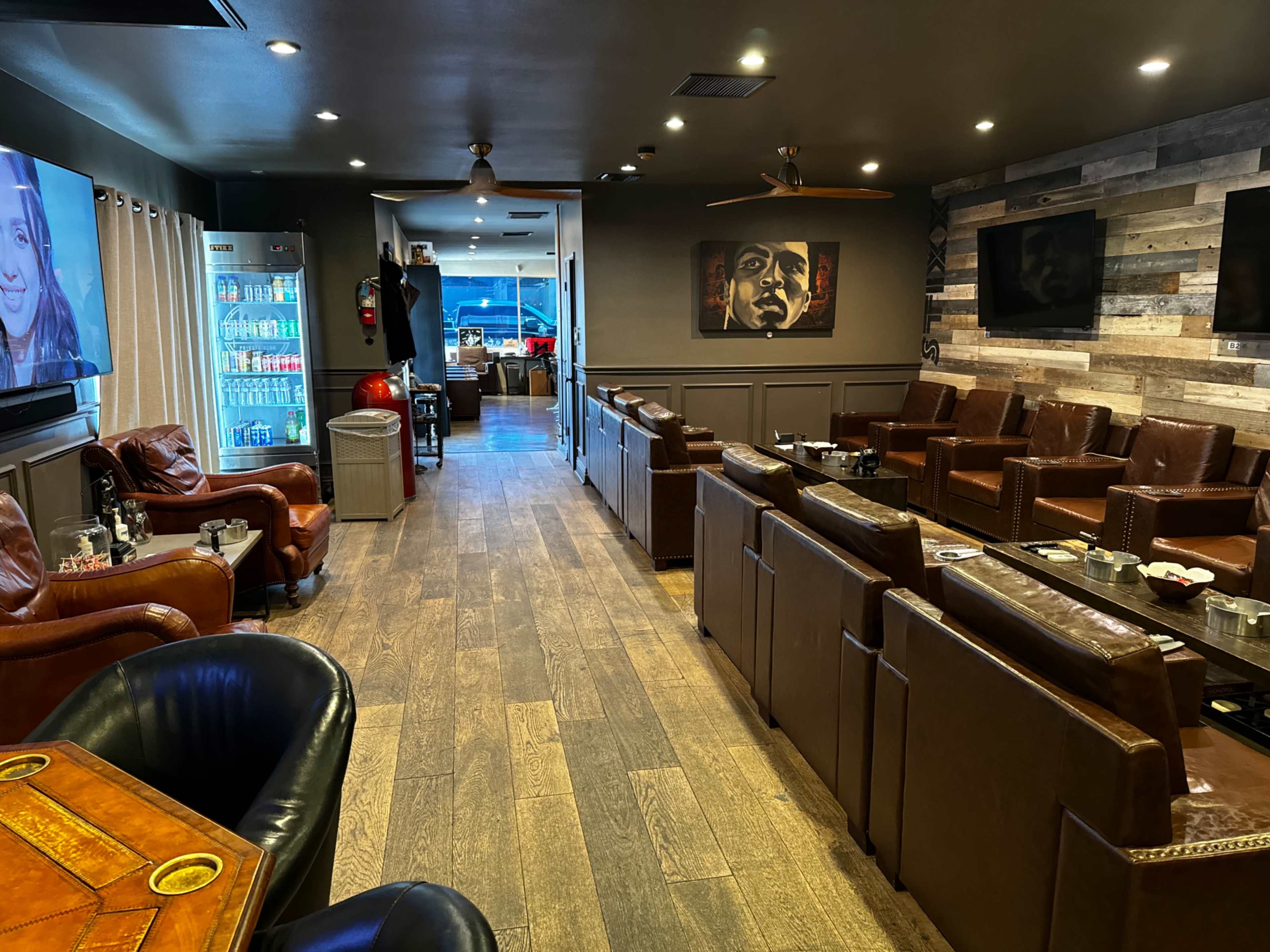 The image features a spacious lounge area with leather seating, wooden flooring, and a variety of artwork on the walls, along with a visible refreshment station and screen.