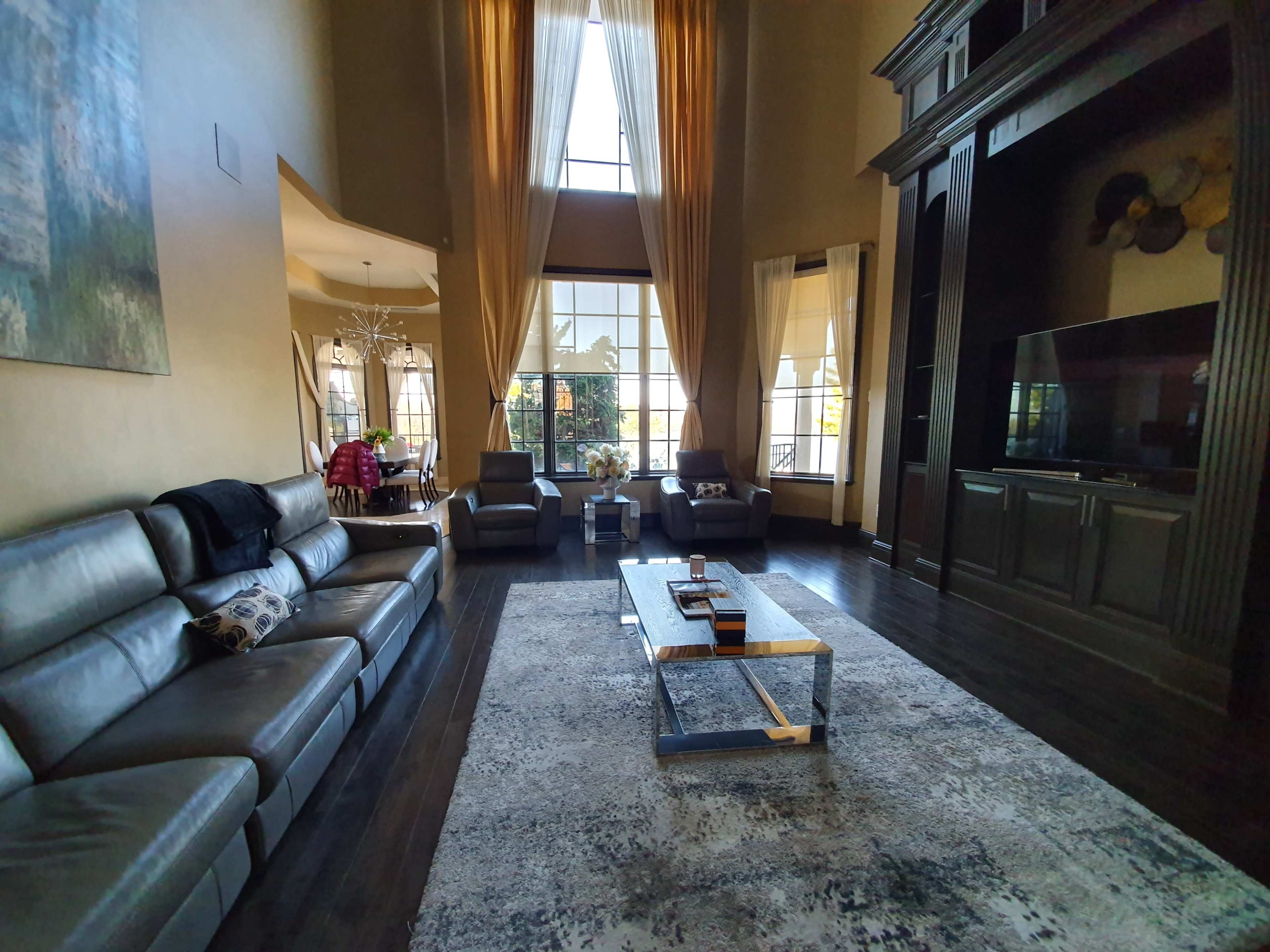 A spacious living room featuring a large window with drapes, a sectional leather sofa, a coffee table, and a television mounted on a dark cabinet.