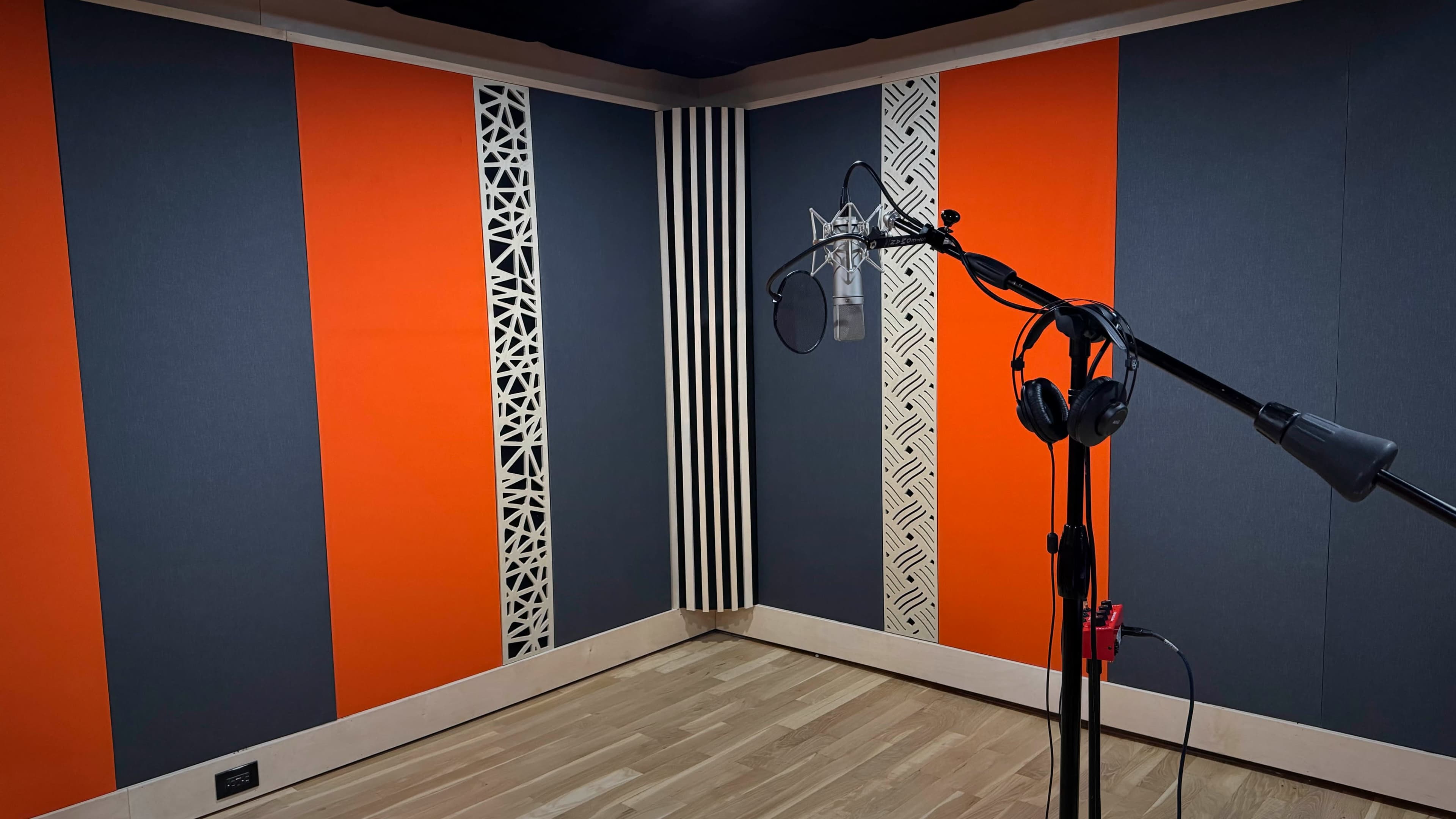 The image shows a soundproof recording studio with orange and gray acoustic panels, a microphone on a stand, and headphones hanging nearby.