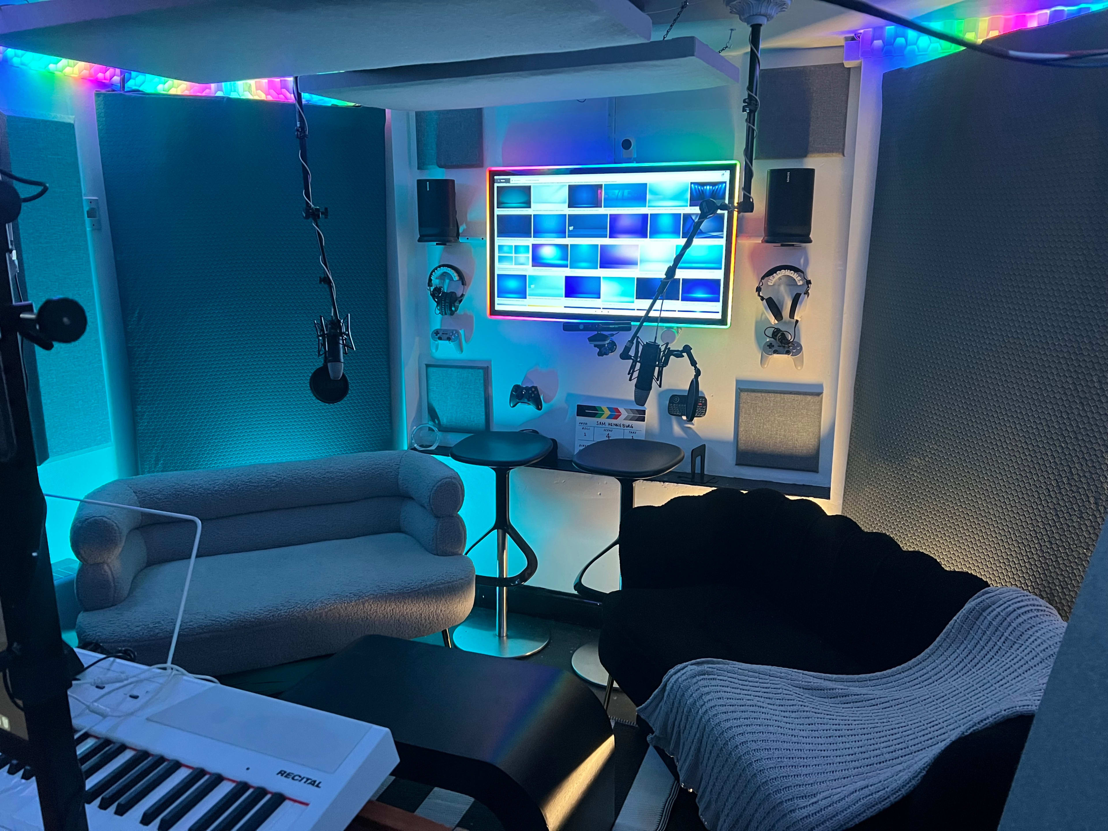 The image shows a modern recording studio setup with two seating areas, a large screen, and various audio and gaming equipment arranged against soundproofed walls.