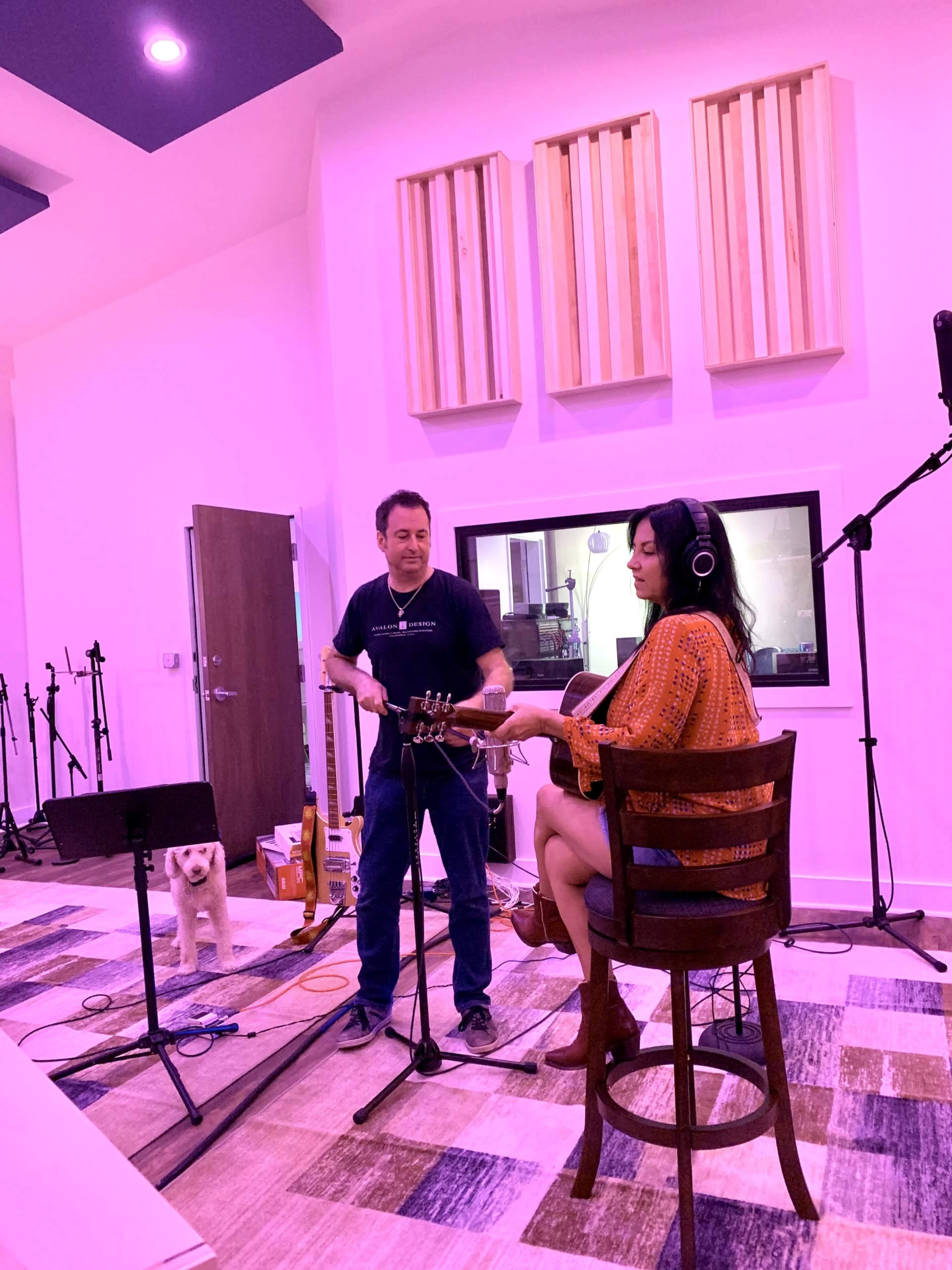 A man stands near a microphone while a woman plays guitar in a recording studio, and a dog watches from the side.