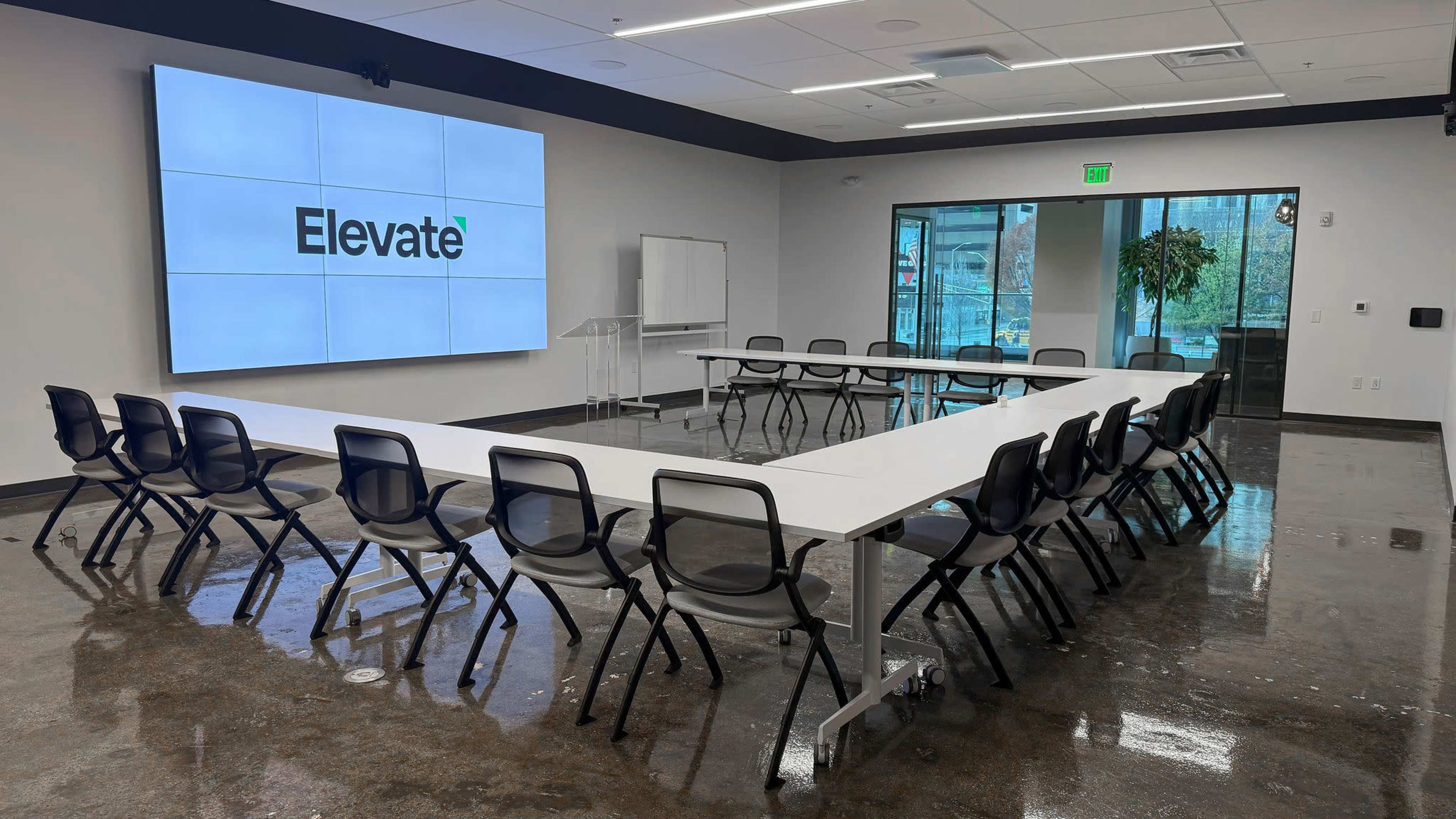 The image shows a modern conference room with a large screen displaying the word "Elevate" and a rectangular arrangement of black chairs around a white table.