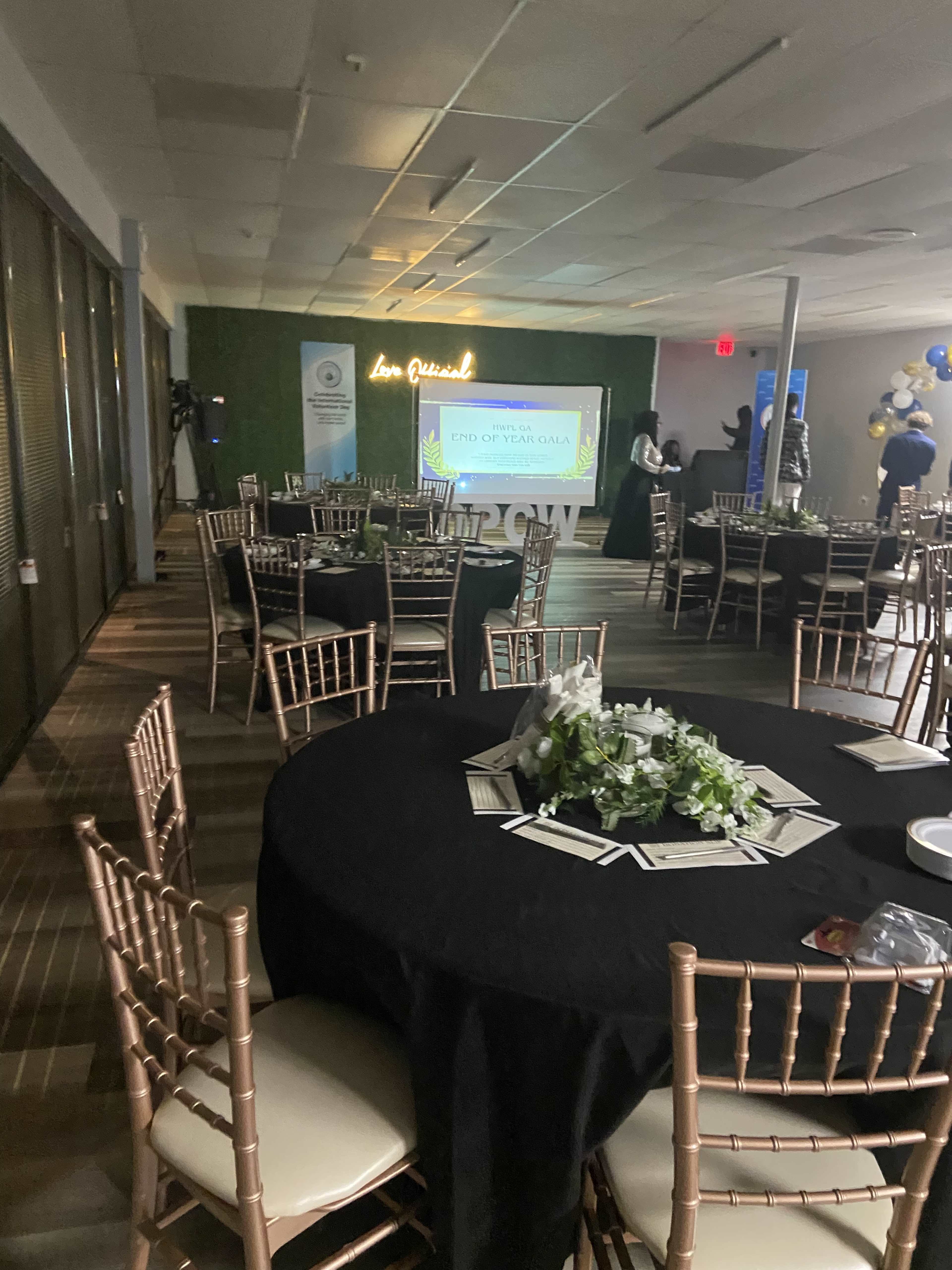 The image shows a banquet hall set up for an event, featuring round tables with black tablecloths, chairs, and a projector screen displaying "END OF YEAR GOALS."