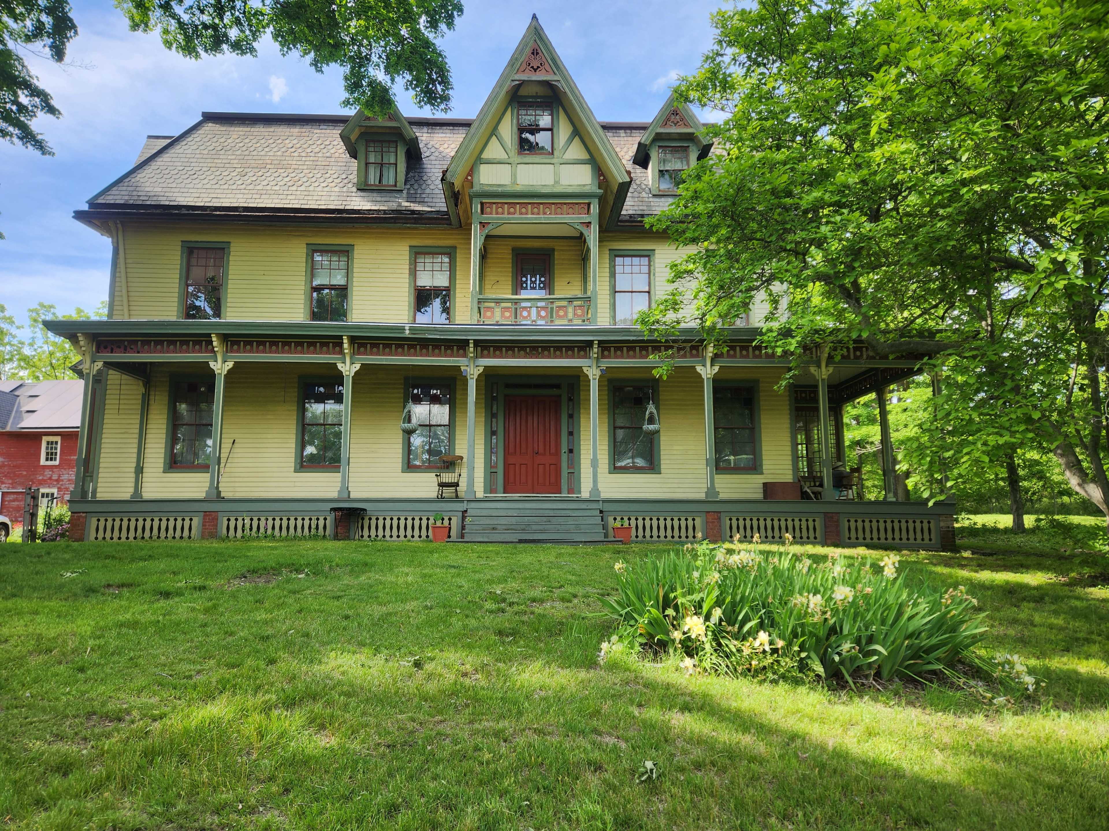 The image shows a large, yellow Victorian-style house with multiple gables and a spacious front porch surrounded by green grass and trees.