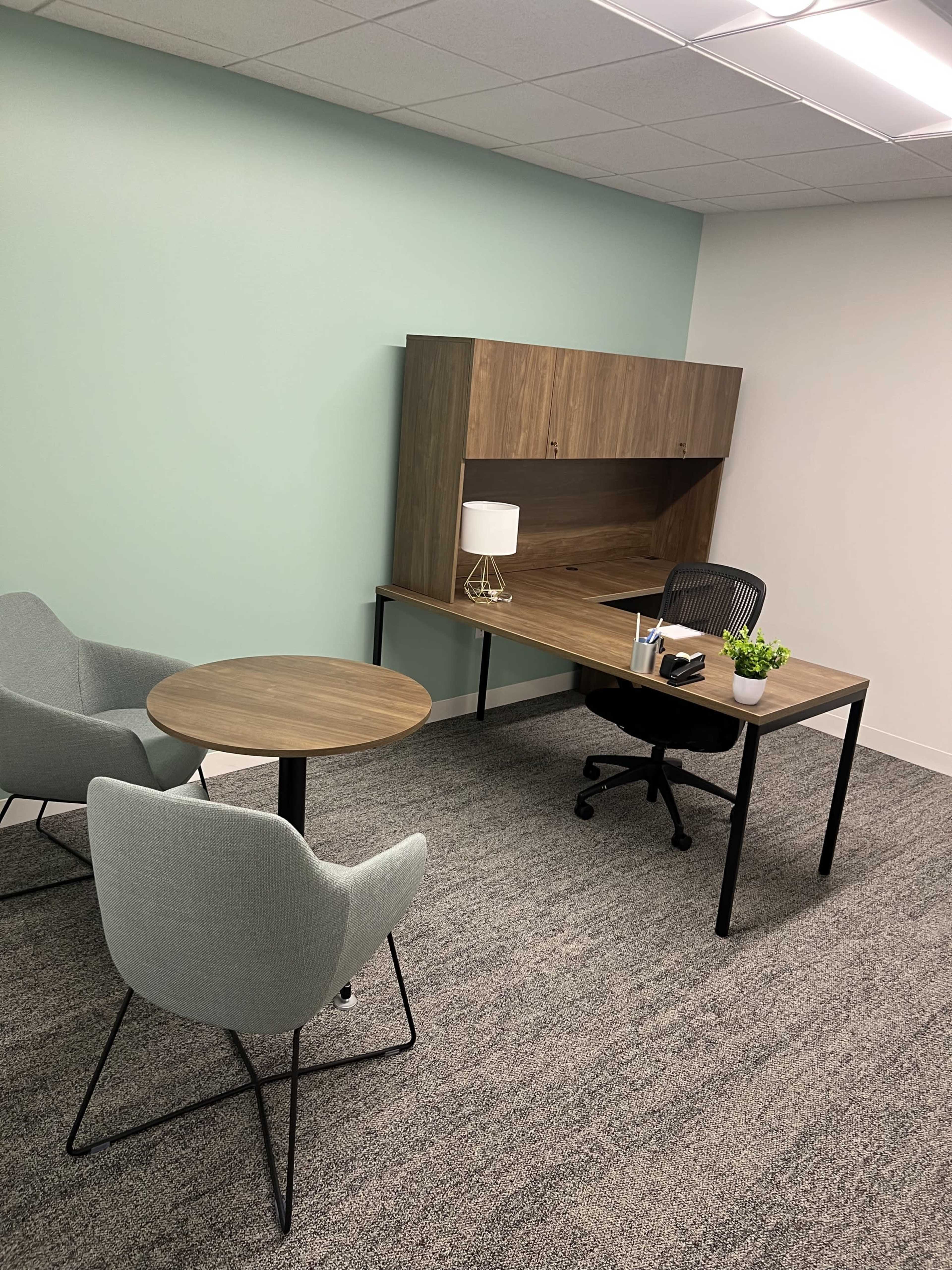 The image shows a small office space with a wooden desk, a chair, a table, and two armchairs, all set against a light green wall.