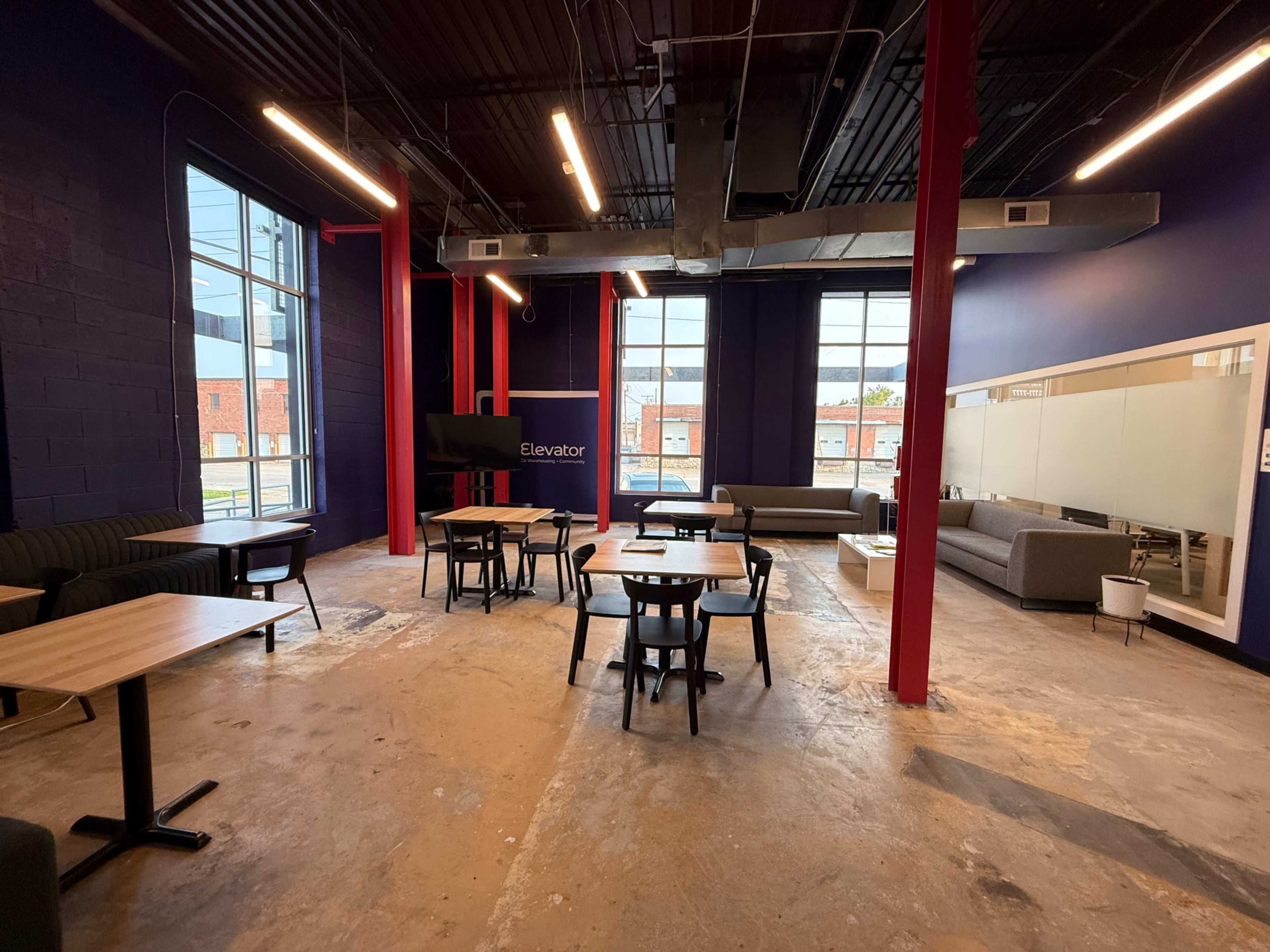 The image shows a spacious, partially furnished lounge area with wooden tables and chairs, large windows, and exposed industrial elements.
