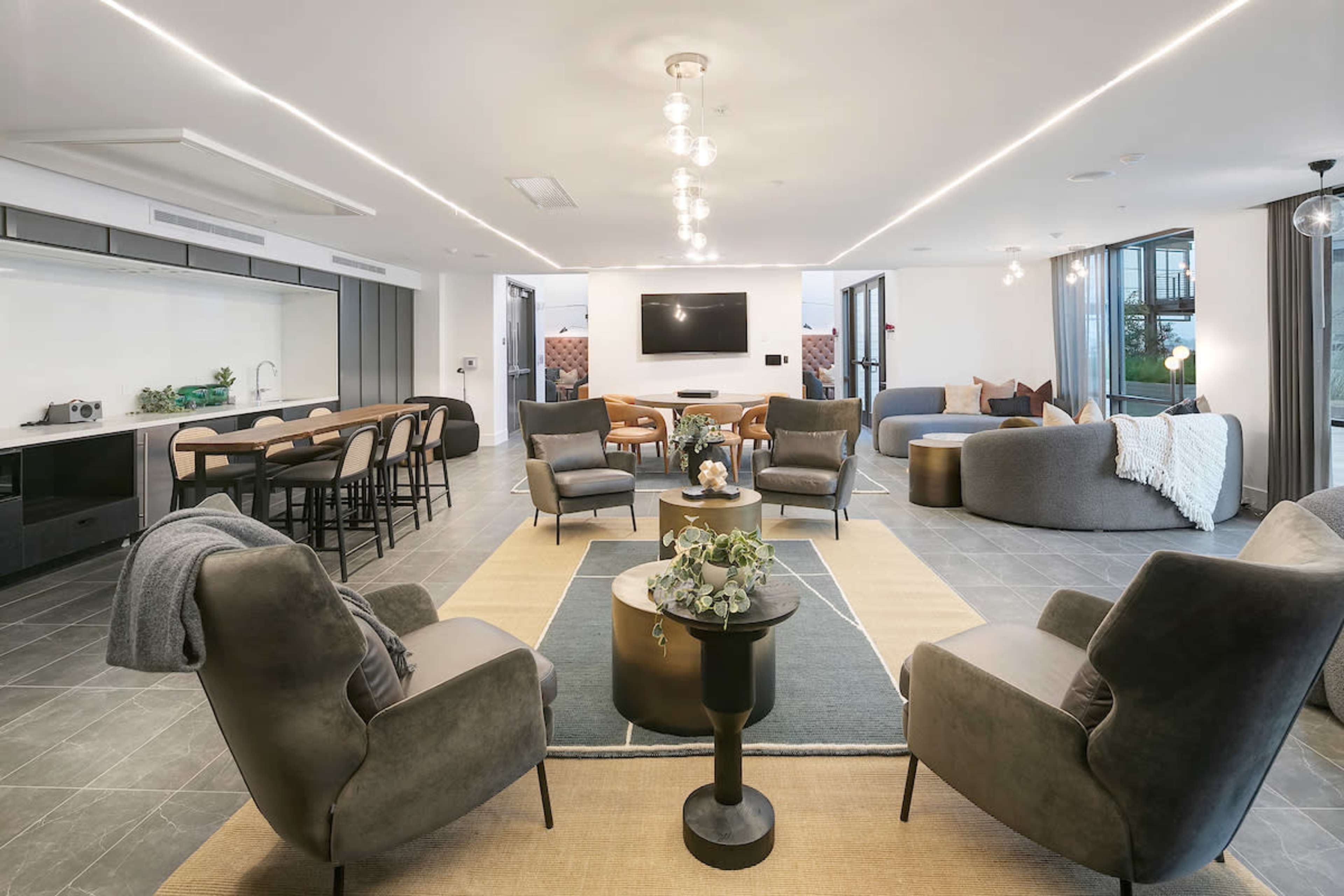 A modern living area featuring a combination of plush seating, a central coffee table, and a kitchen area with bar stools, all set against a backdrop of large windows and a minimalist decor.