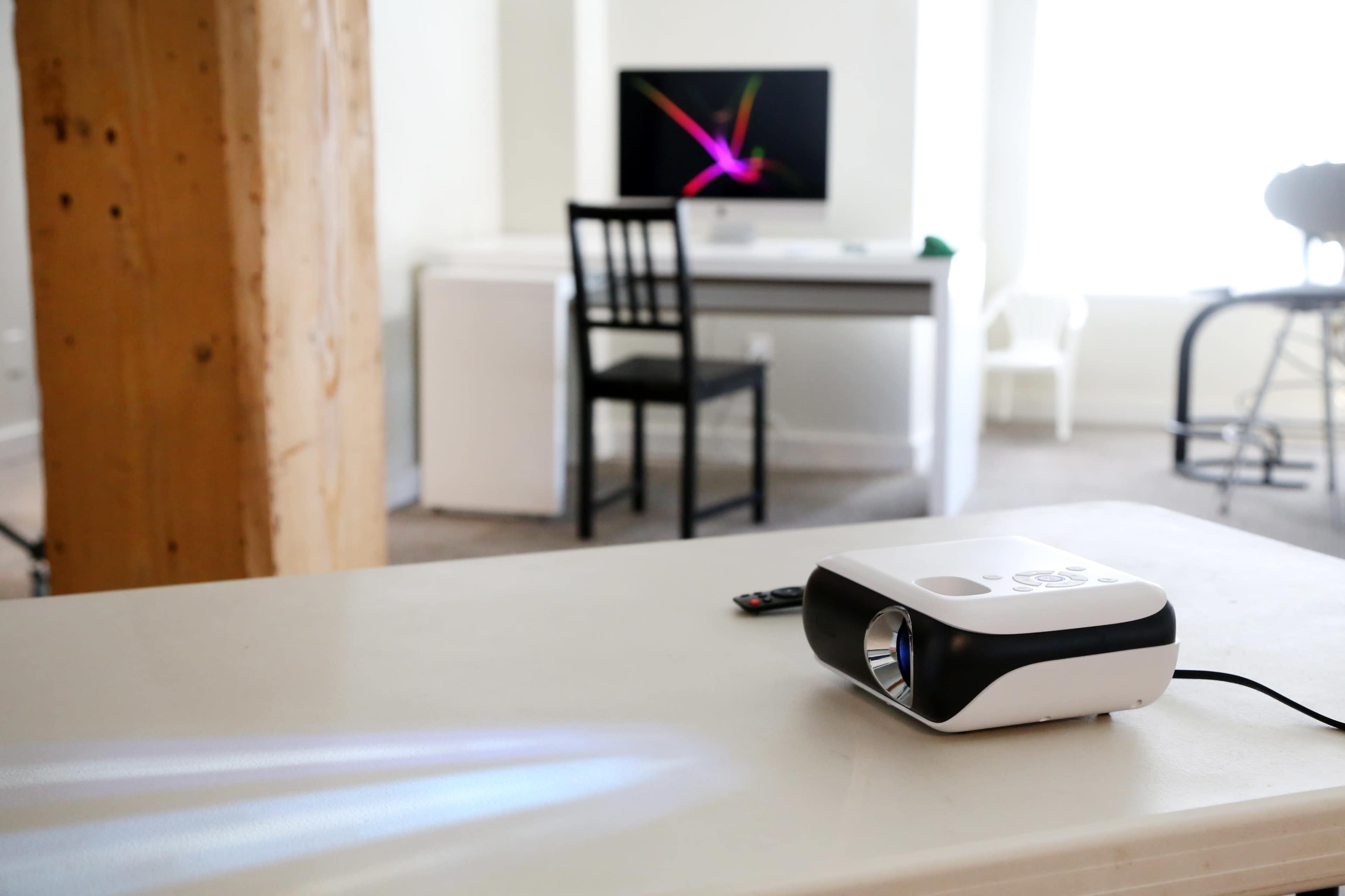 A projector sits on a table, casting light, with a desk and chair visible in the background.