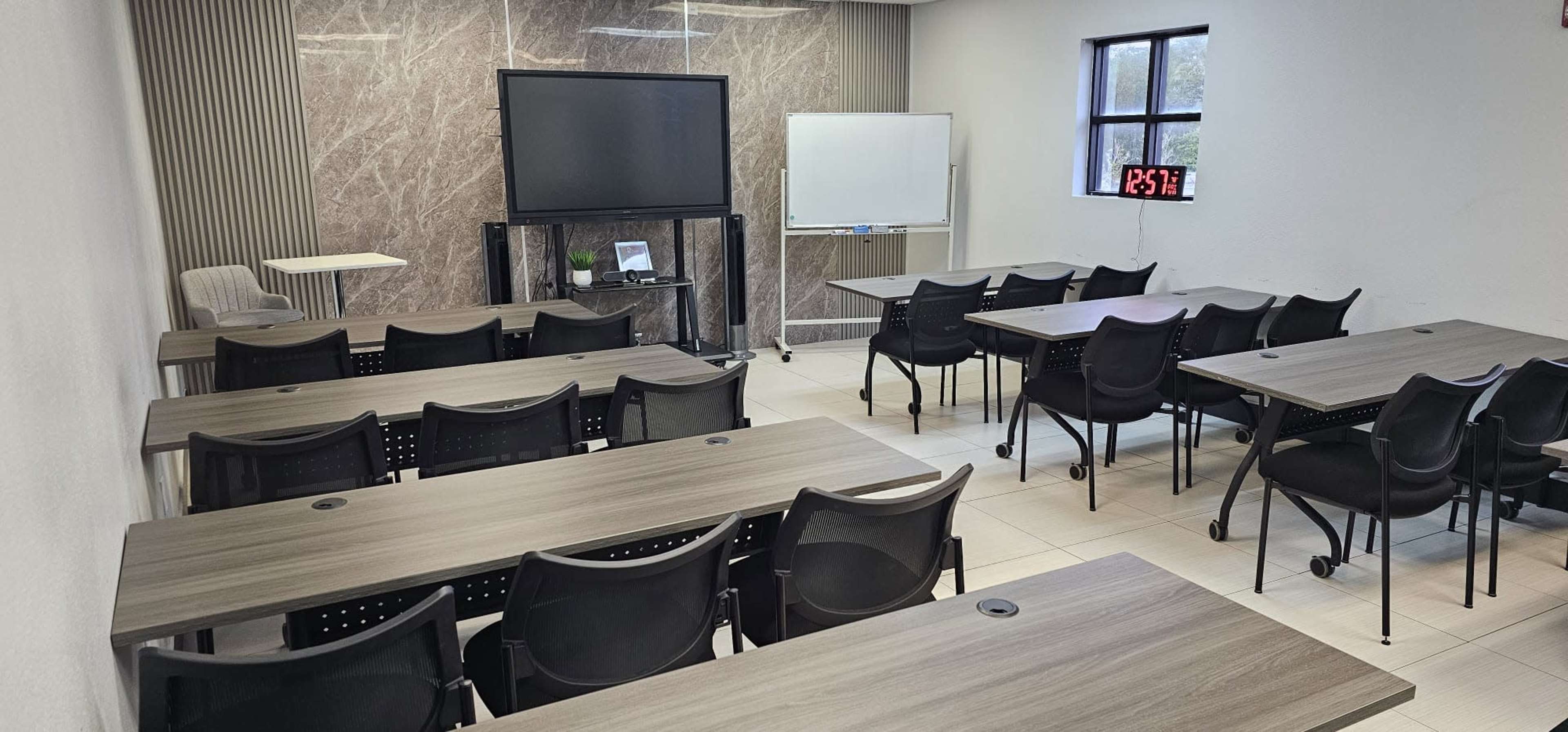 The image shows a modern classroom setup with several rows of tables and chairs, a large screen on a stand, a whiteboard, and a clock displaying the time.