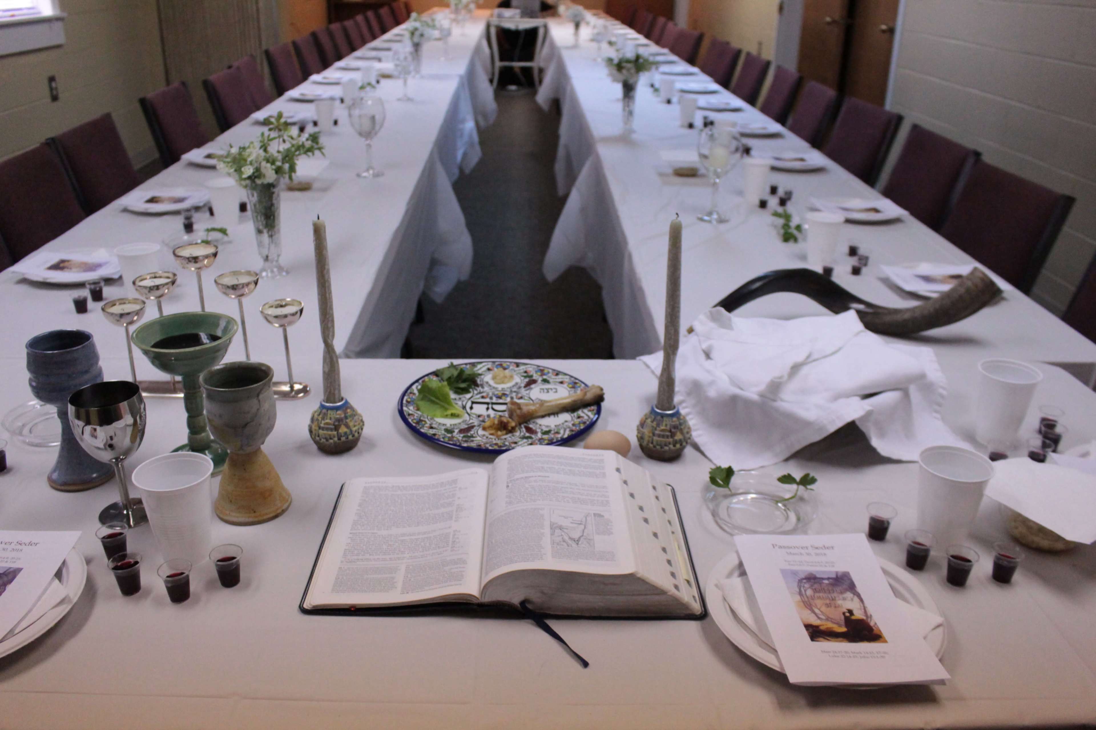 A long dining table is set with white tablecloths, glassware, and a variety of ceremonial items, including a large open book and small cups arranged along the center.