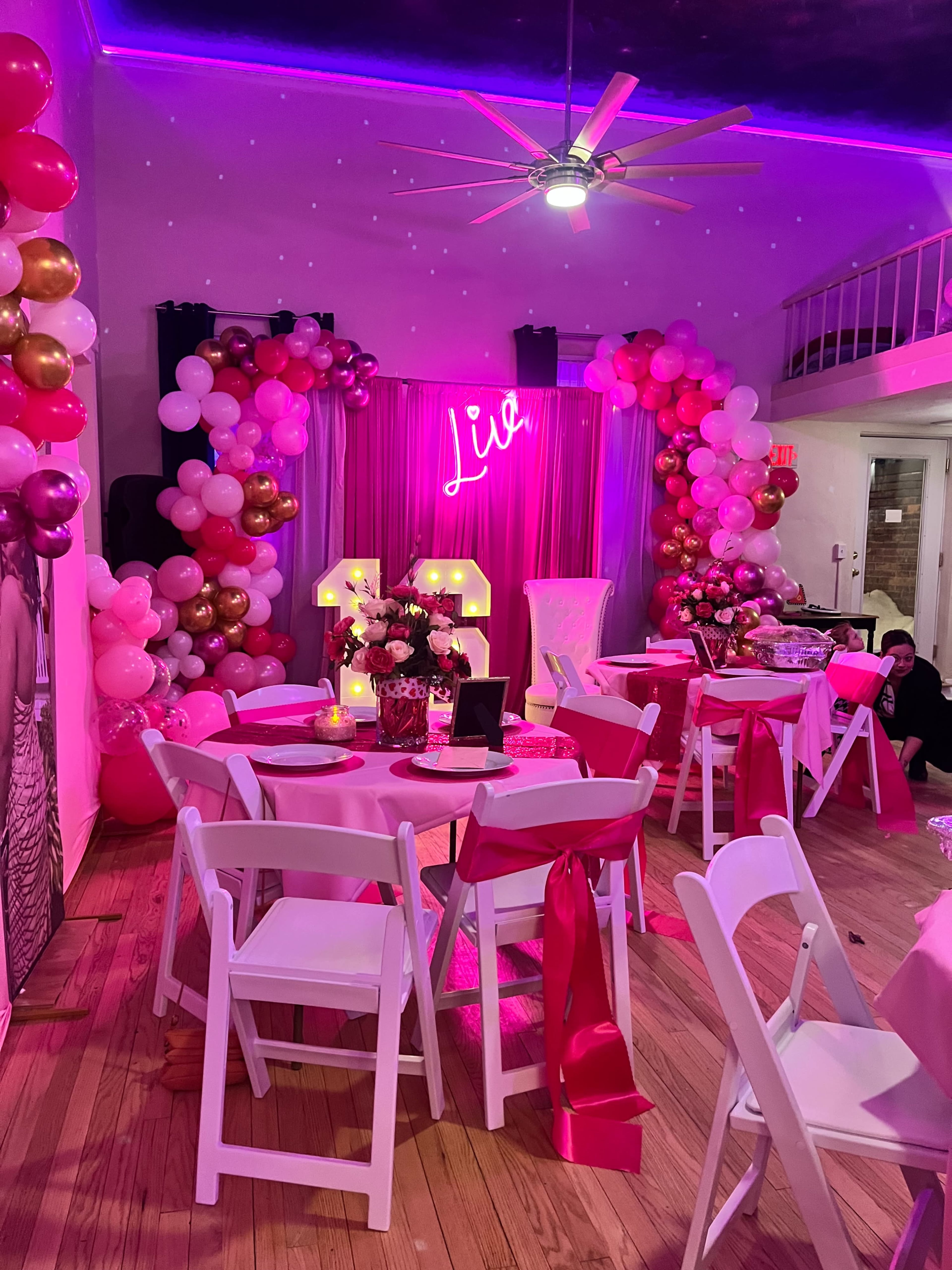 The image shows a decorated event space featuring pink and gold balloons, round tables with white chairs, and a neon sign that reads "Live."