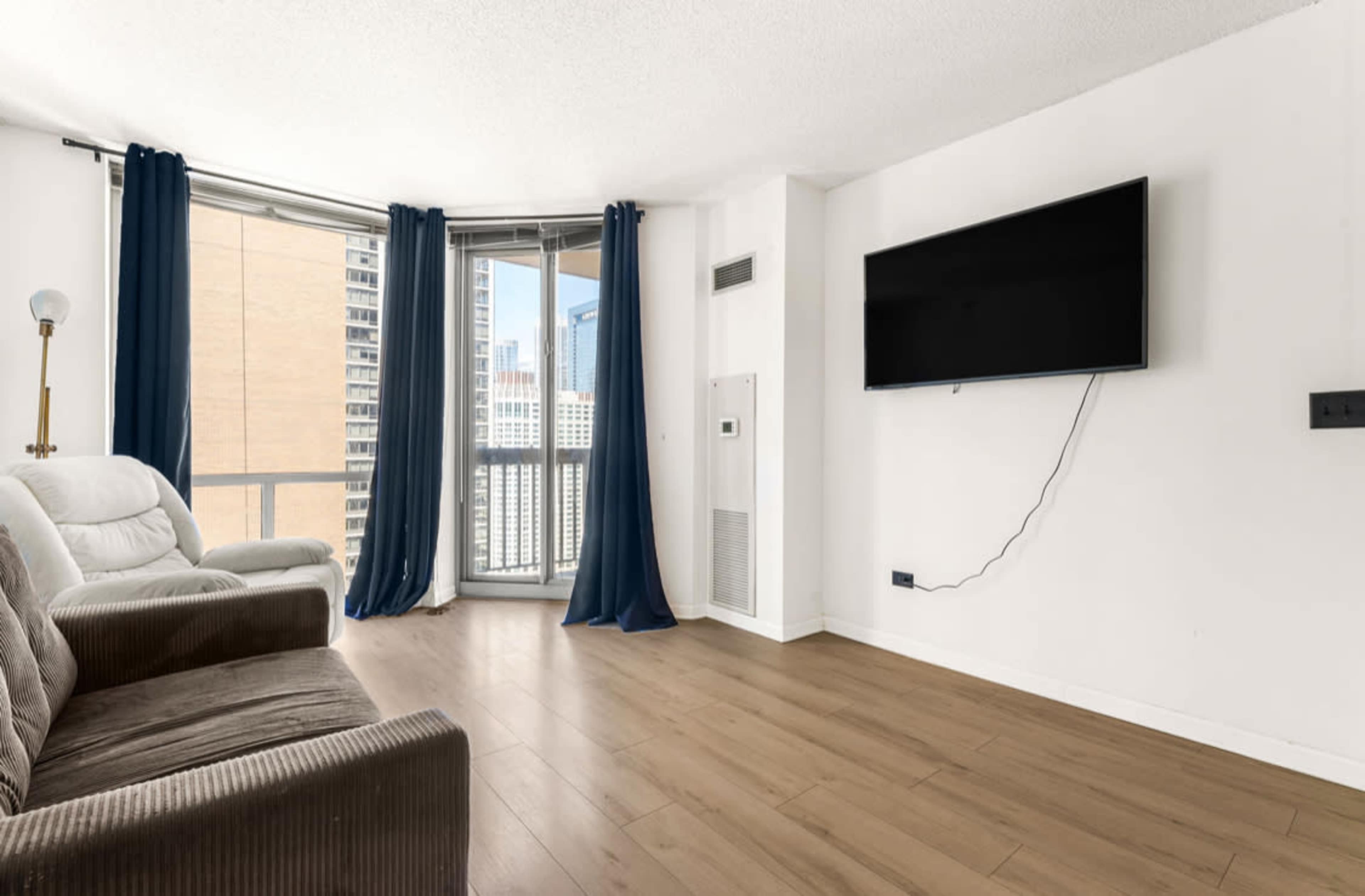 The image shows a modern living room with a large window, a flat-screen TV mounted on the wall, a recliner chair, and wood flooring.