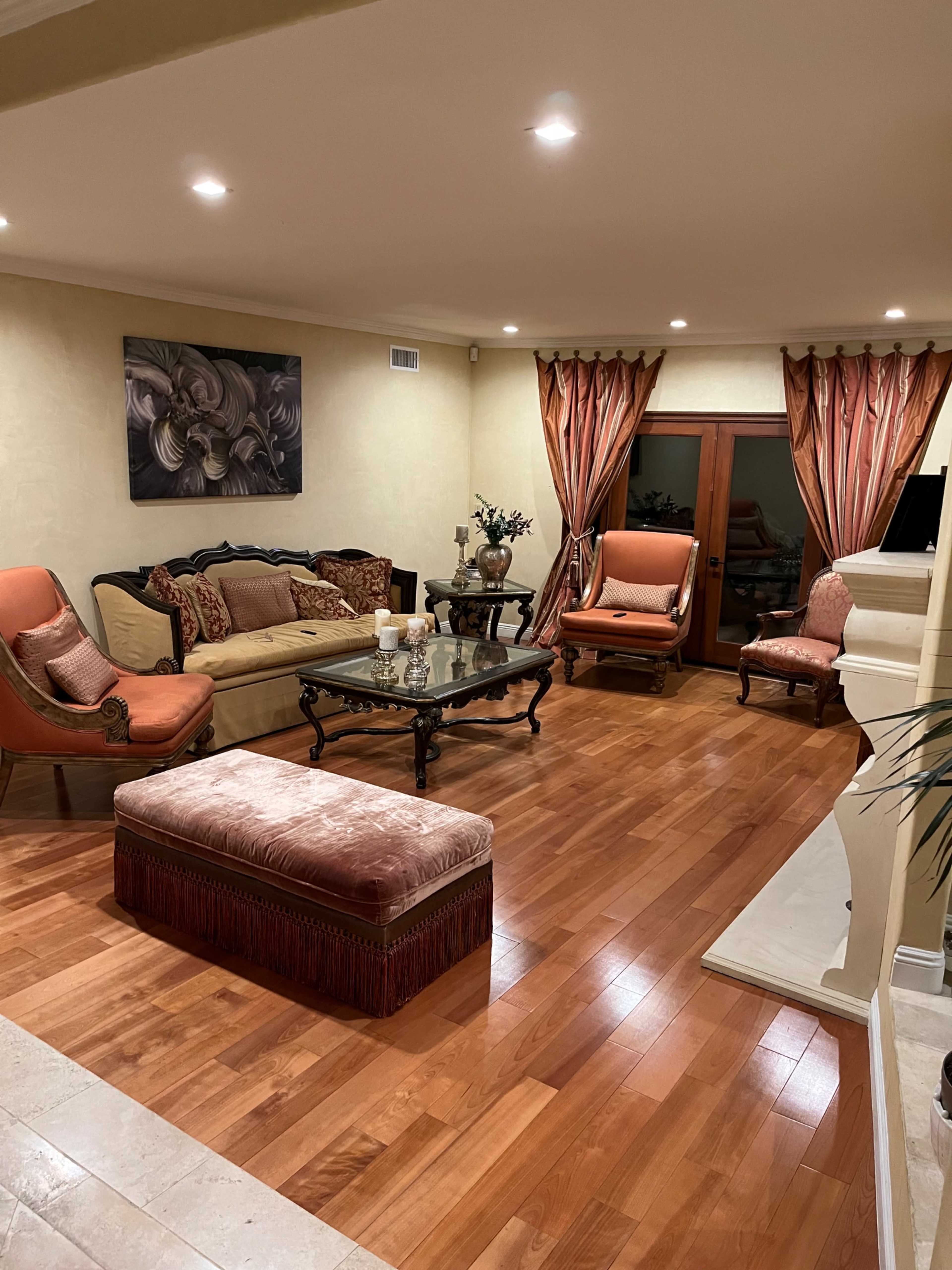 The living room features a glass coffee table surrounded by upholstered furniture and wooden flooring, complemented by large windows with draped curtains.