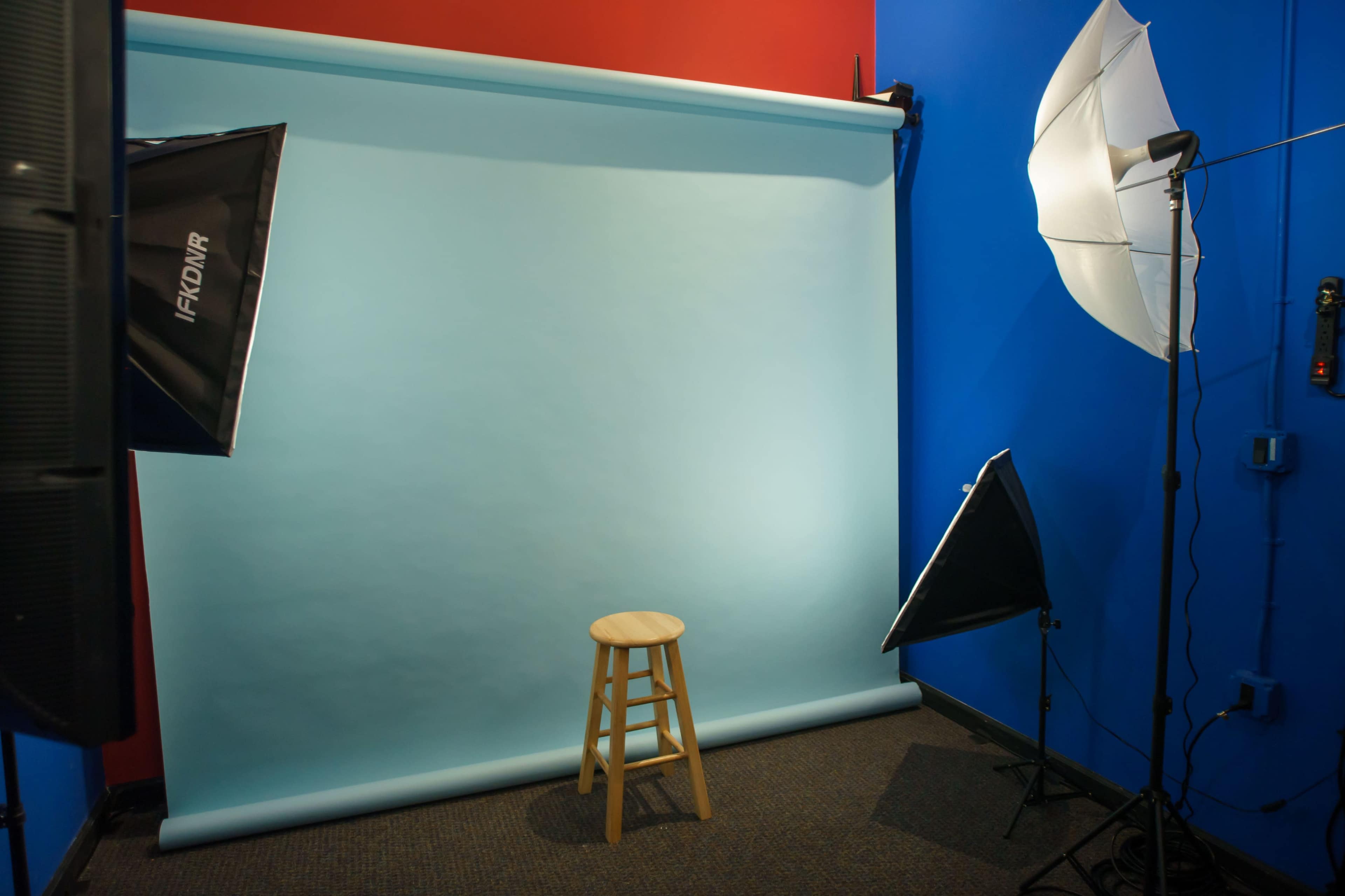 A photography setup features a blue backdrop with a wooden stool in front, flanked by softbox lights on either side.