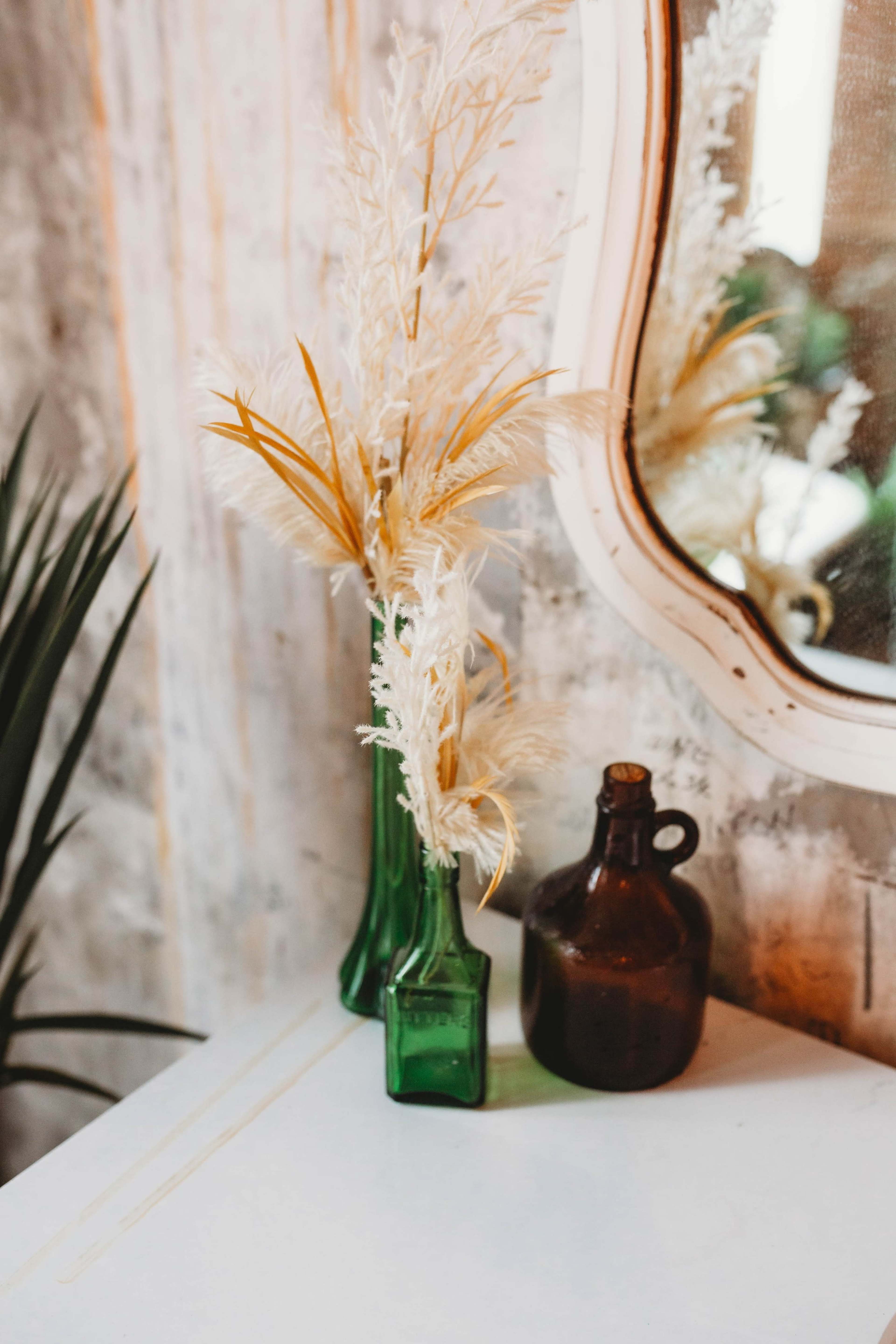 A cluster of two green glass vases filled with dried plants sits on a white surface beside a brown ceramic jug, reflected in a nearby mirror.