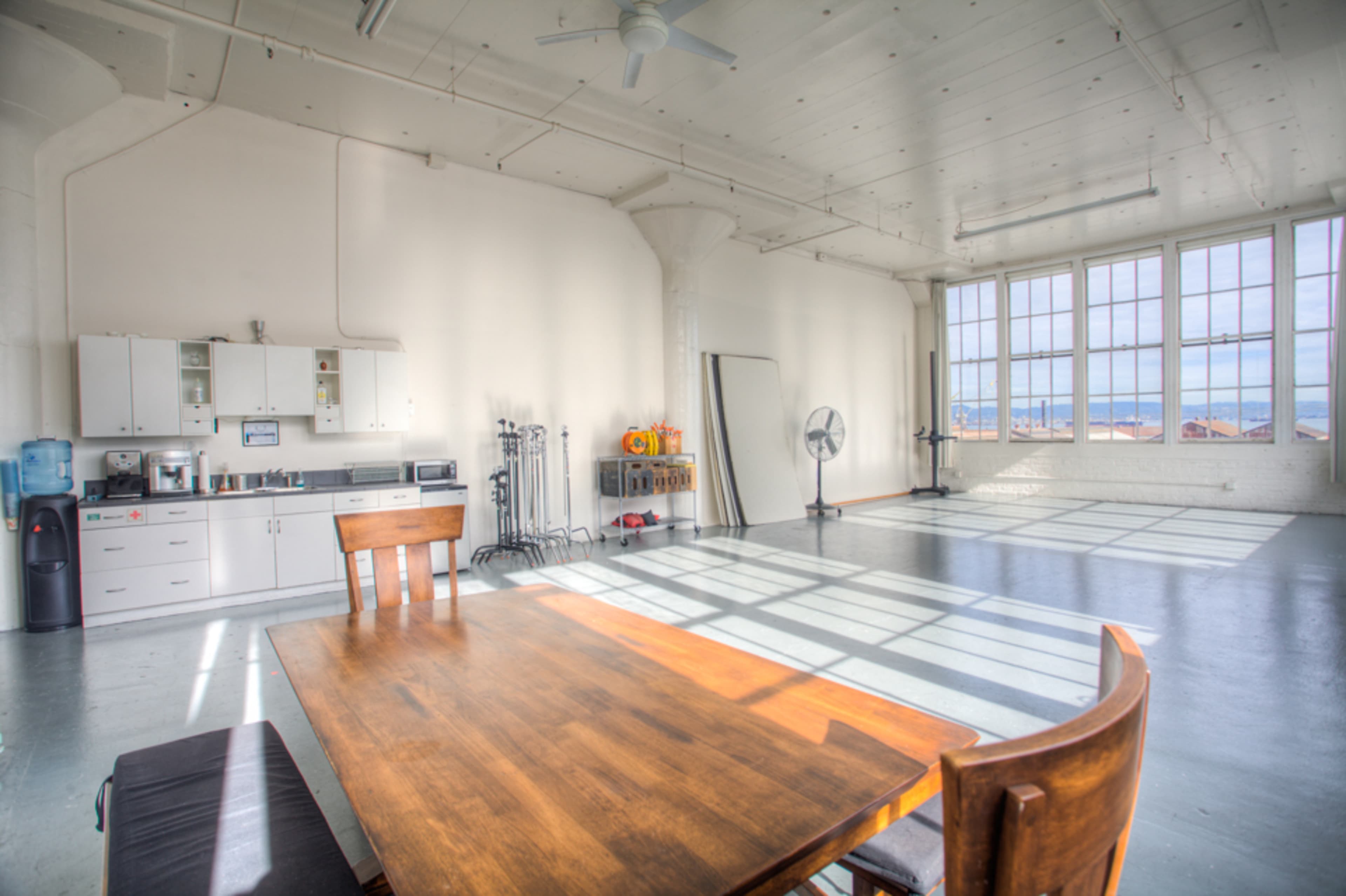 A spacious, brightly lit room features a wooden table, chairs, a kitchenette, and large windows casting shadows on the floor.