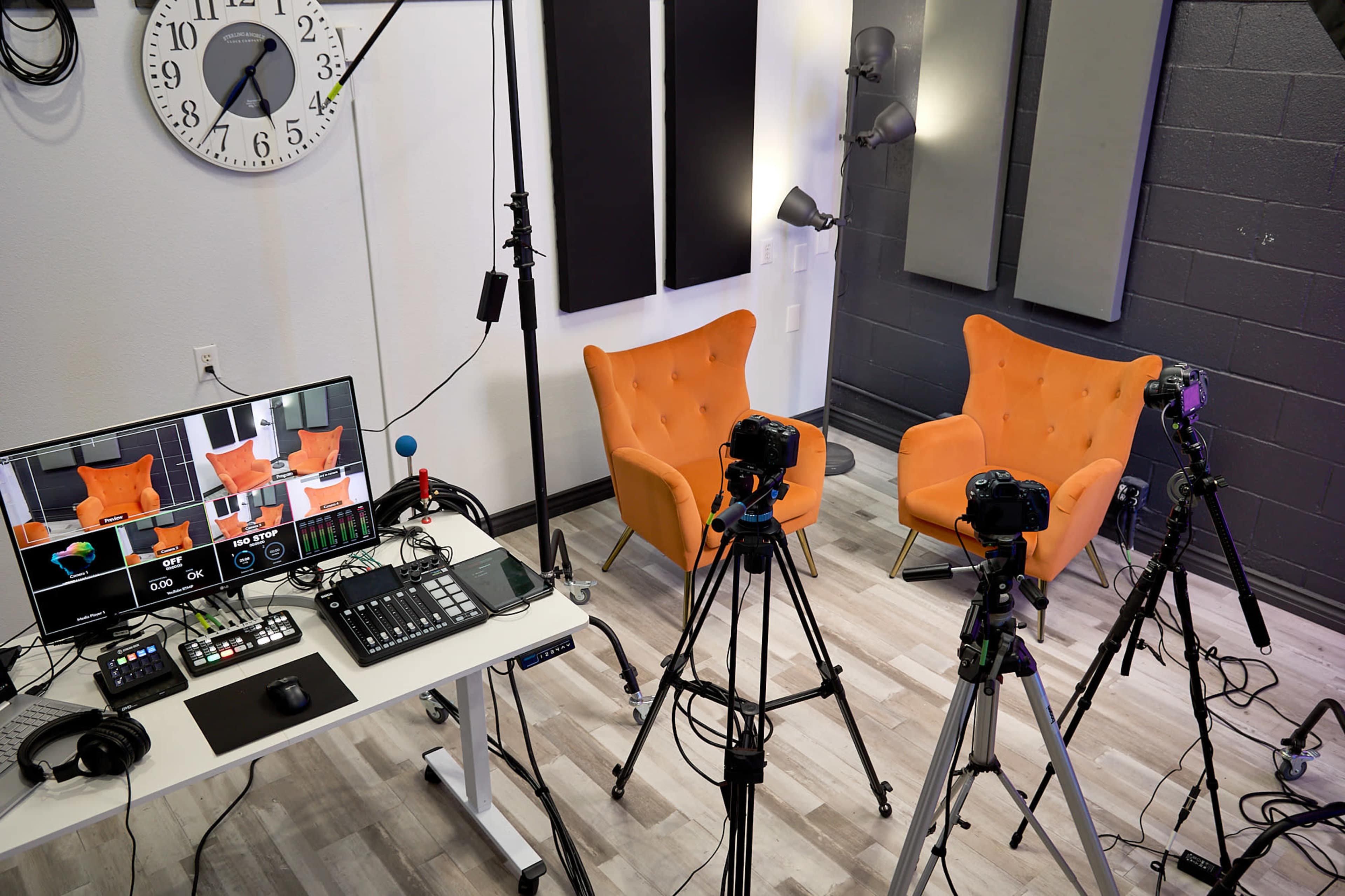 A recording studio setup featuring two orange chairs, multiple cameras on tripods, and a control panel with screens and audio equipment.