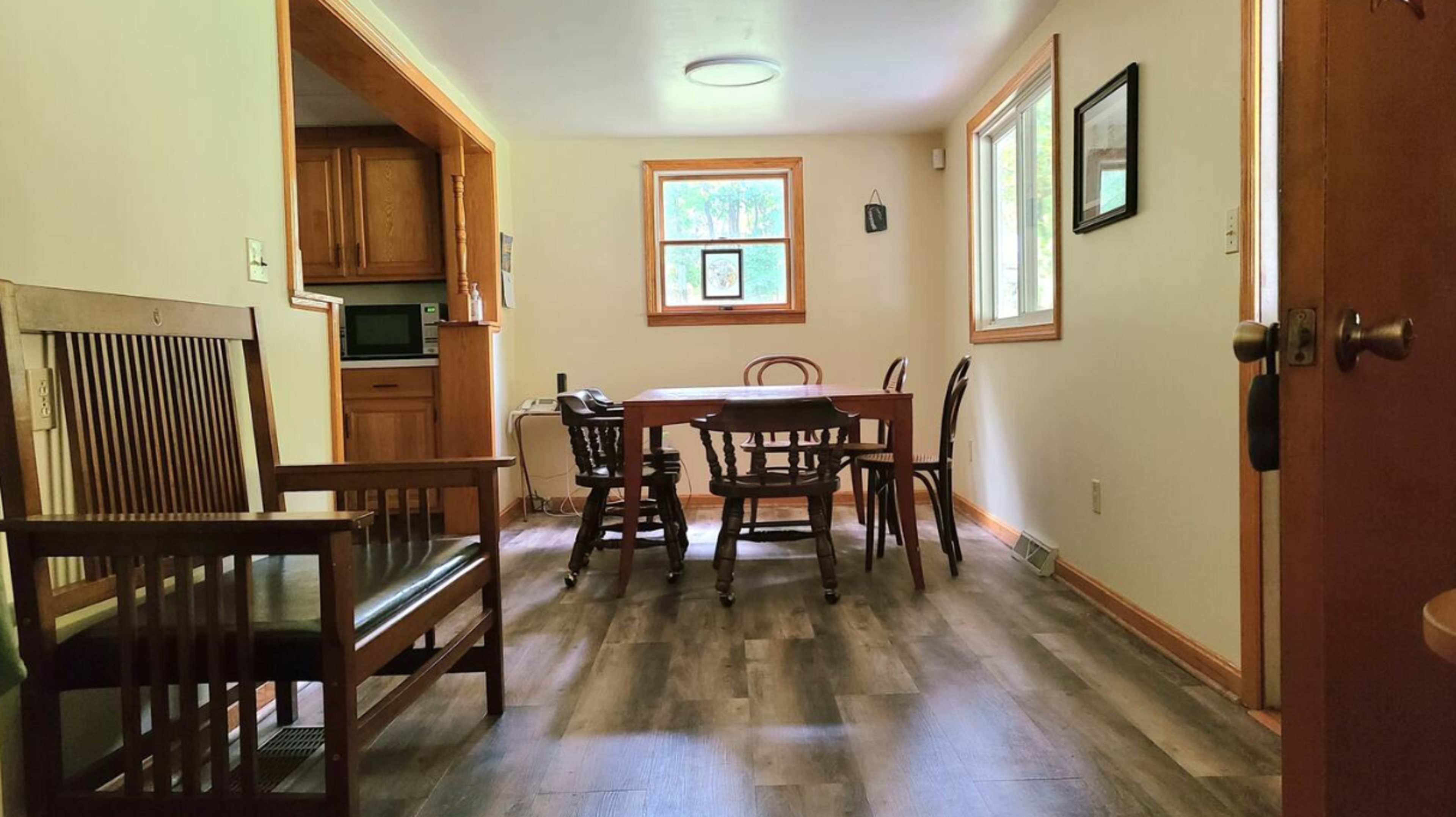 A dining area with a wooden table surrounded by chairs and a small kitchenette visible in the background.