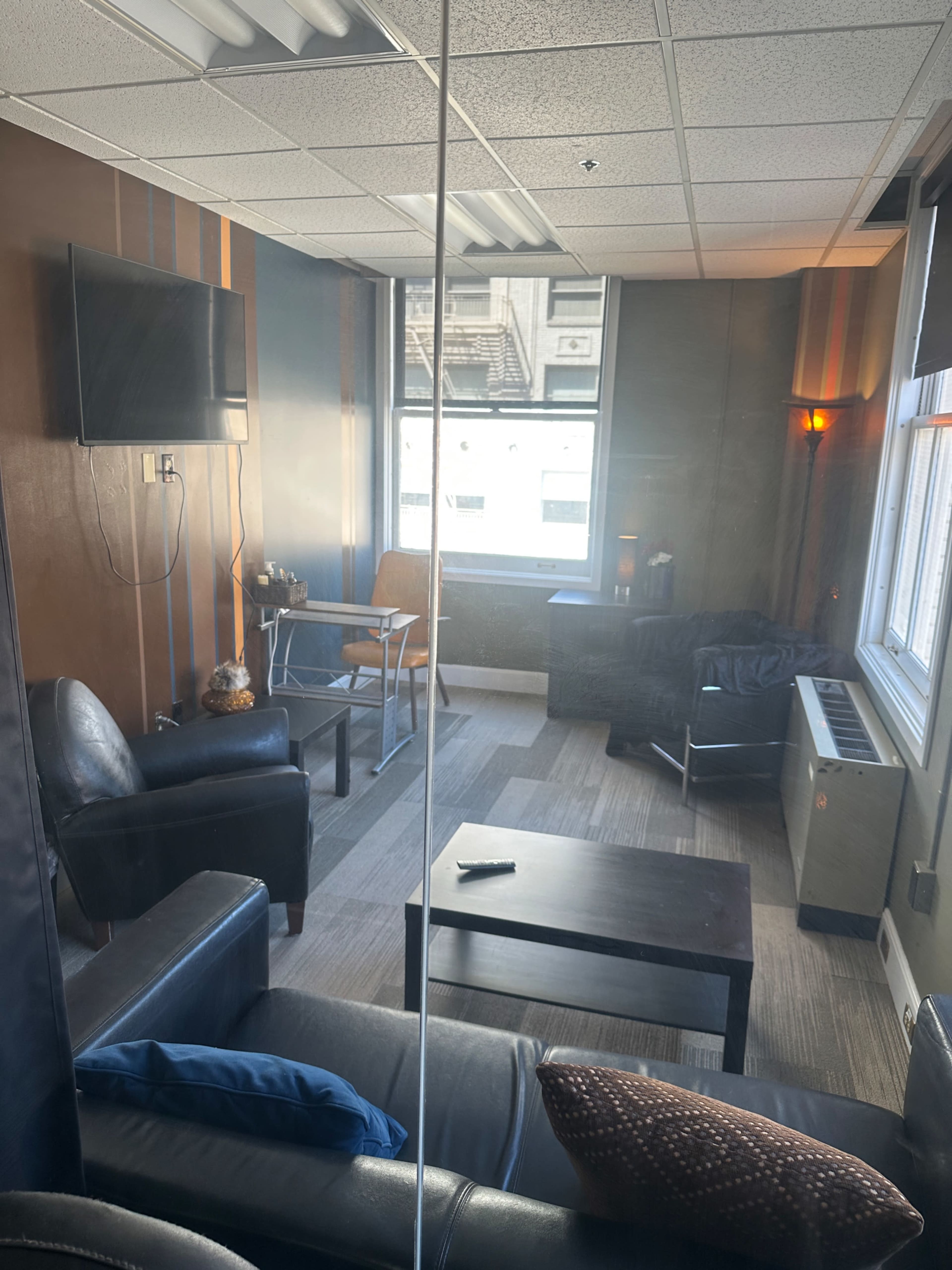 The image shows a small, well-lit office room with a television, two black leather chairs, a table, and a window overlooking a building.