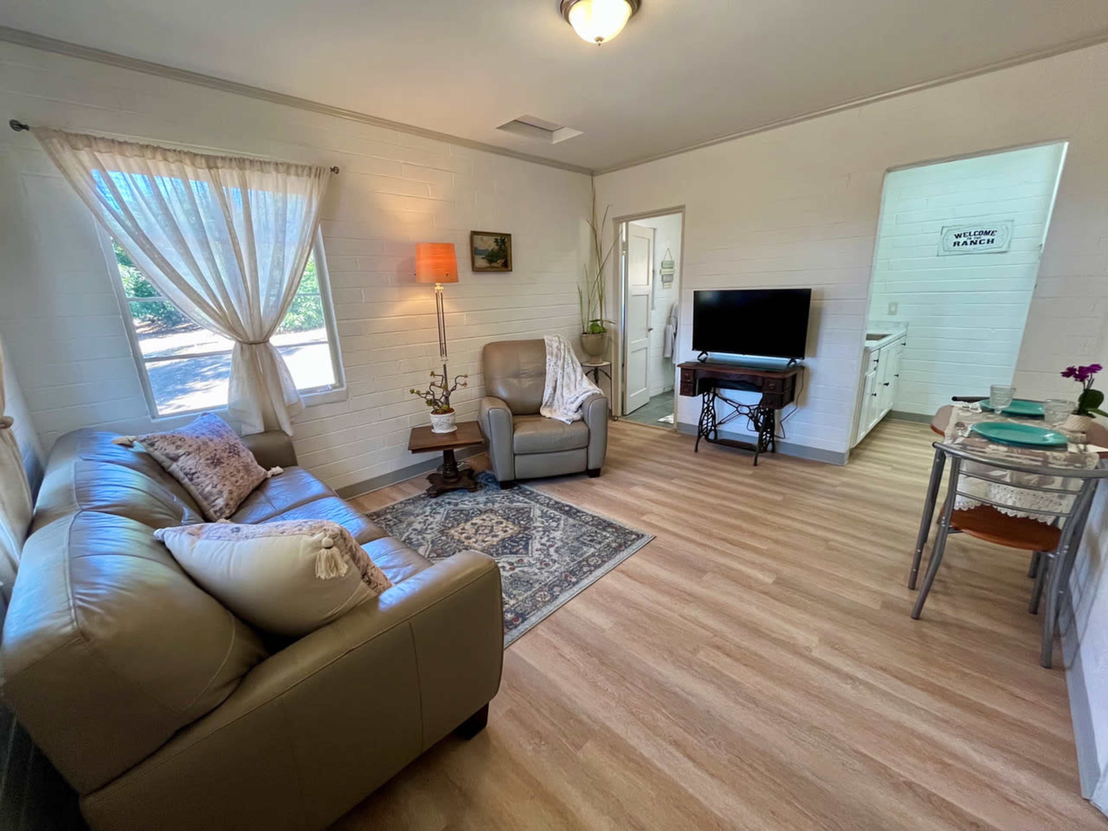The image shows a cozy living room with a beige couch, a small wooden table, a television, and a window with light curtains, plus a door leading to another room.