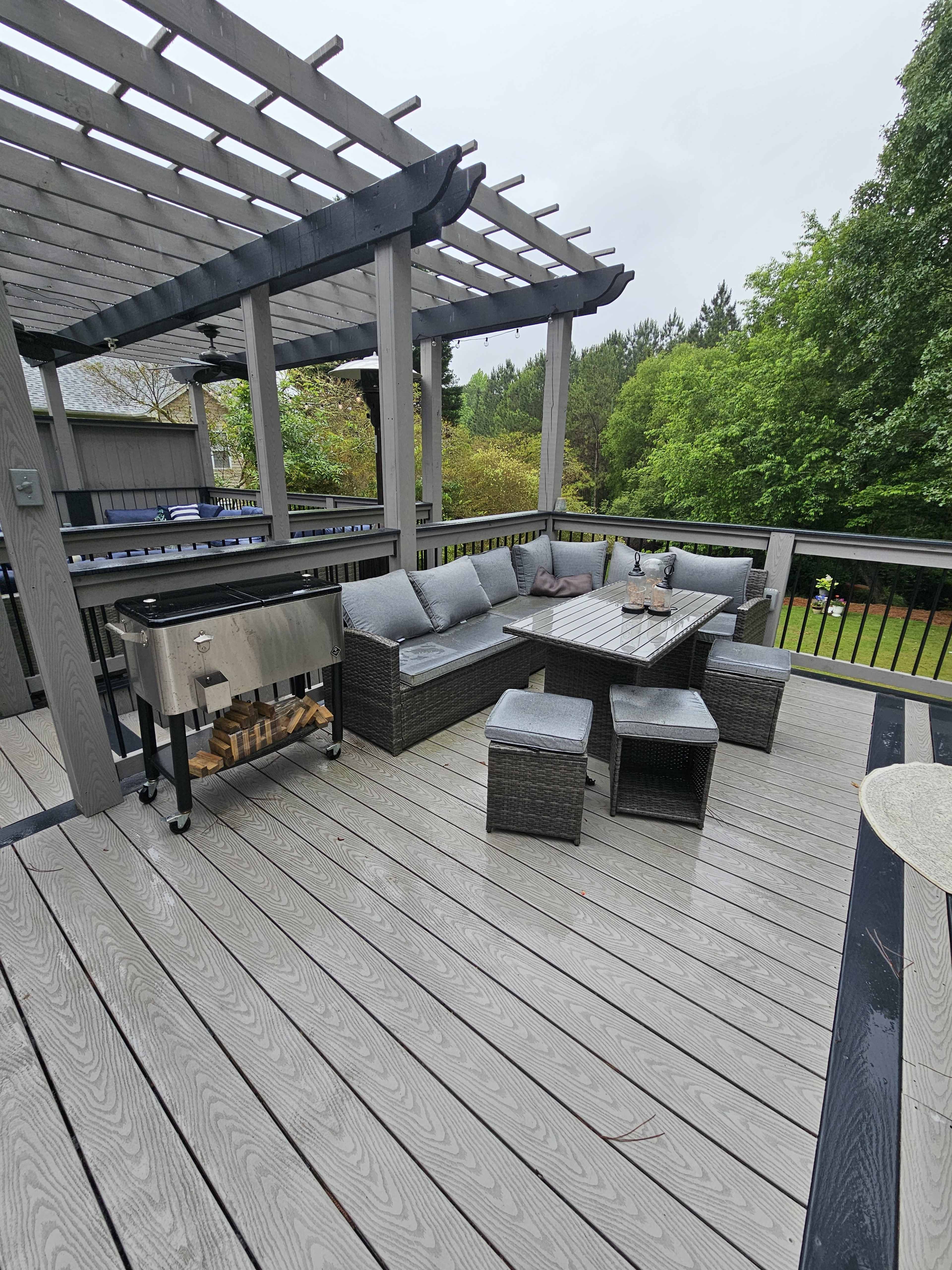 A covered outdoor deck featuring a sectional sofa, a table, and a grill, surrounded by greenery.