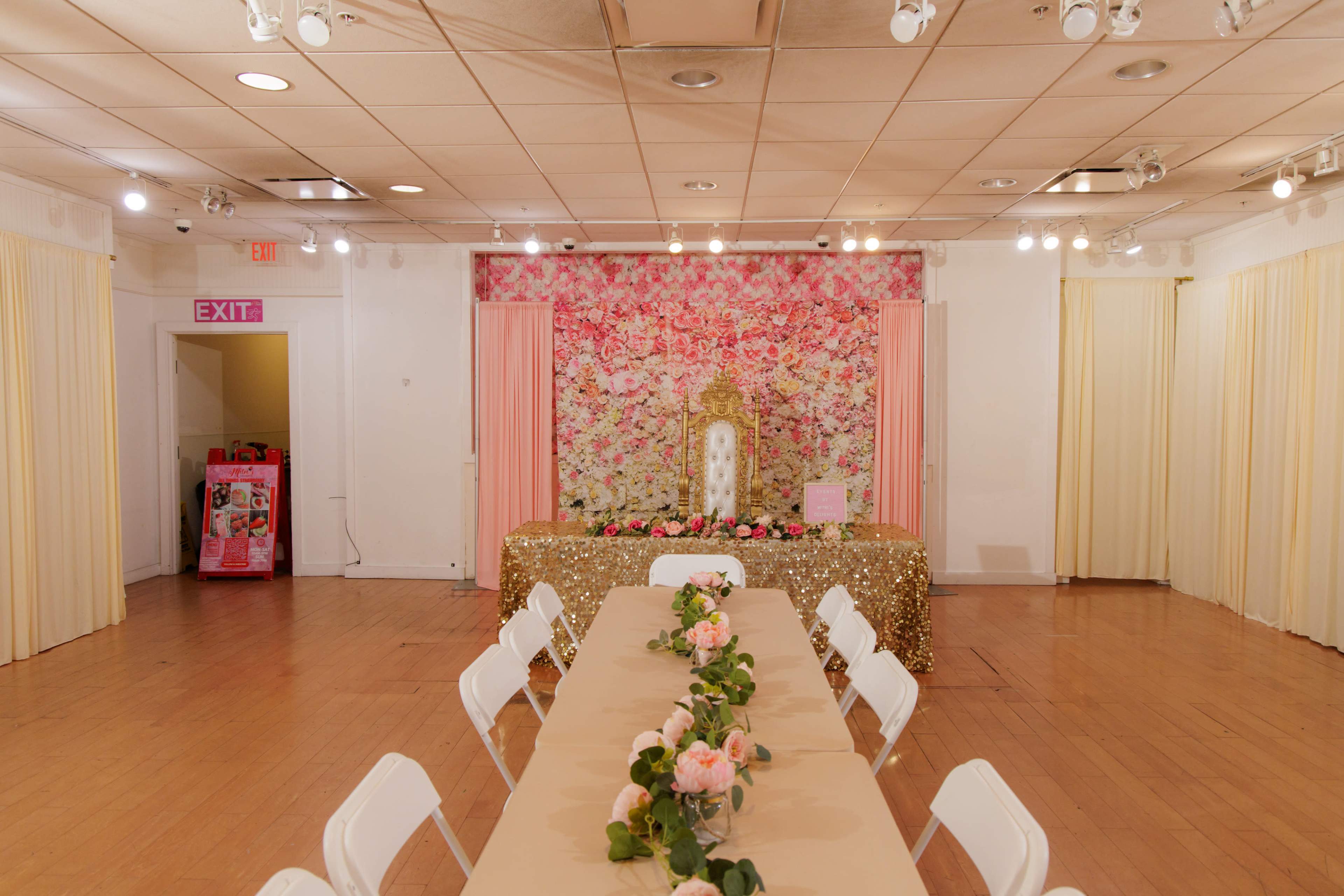 The image shows a banquet hall set for an event, featuring a long table with floral décor, gold sequin decorations on the table, and a floral backdrop with a throne-like chair.